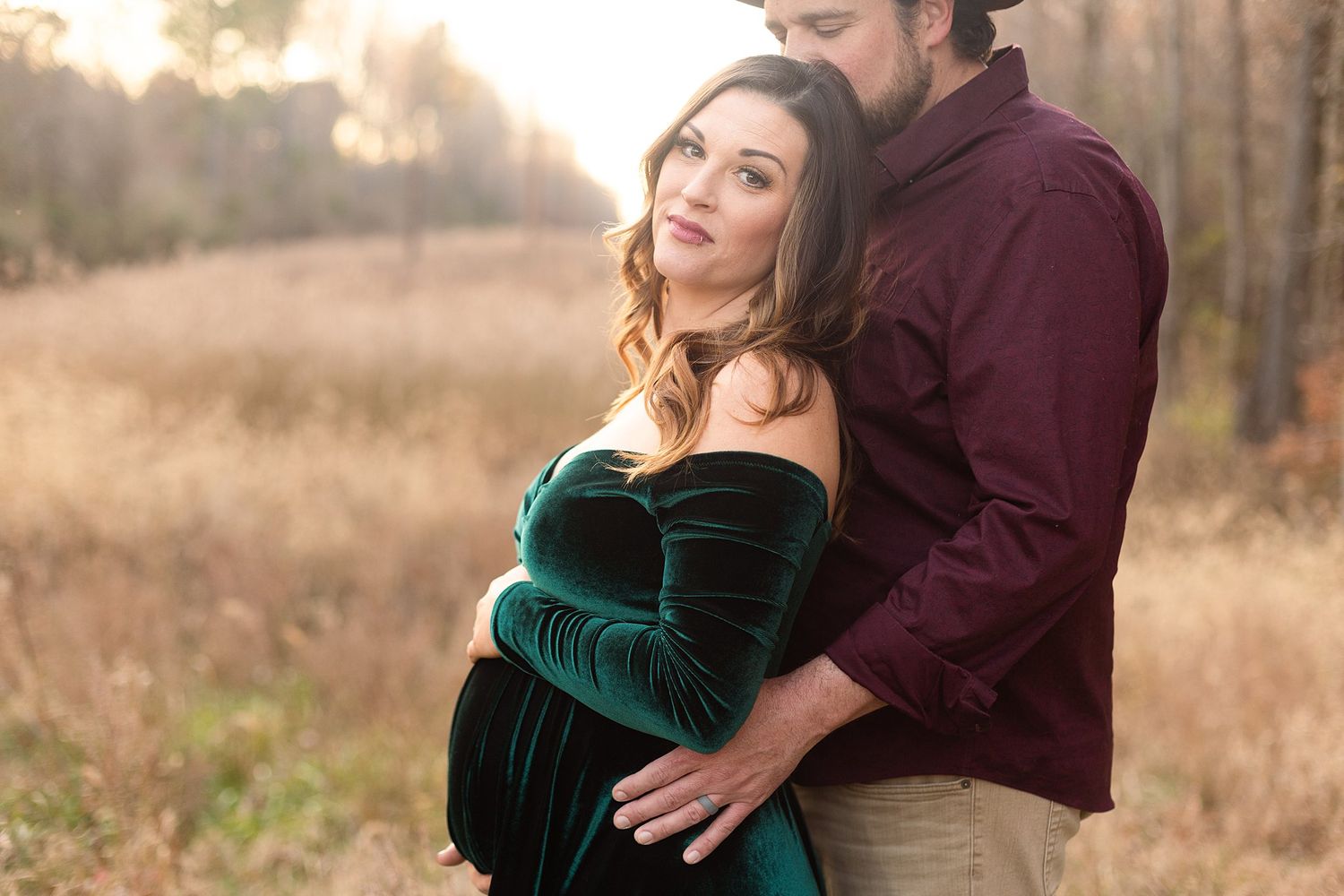 Close-up maternity portrait of couple in burgundy and emerald velvet against golden field.