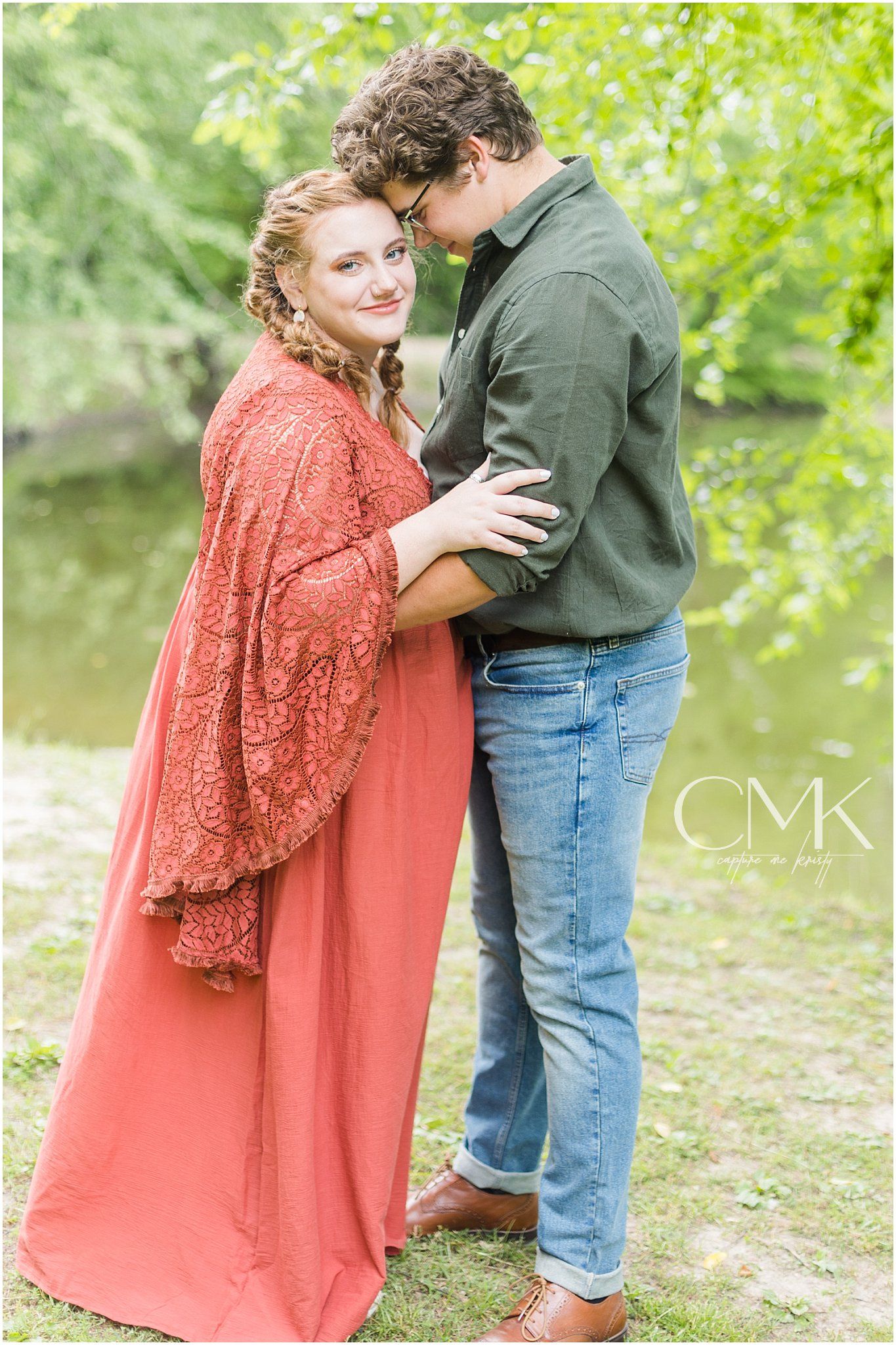A couple embraces outdoors in a romantic engagement photo with a natural green backdrop by a river.