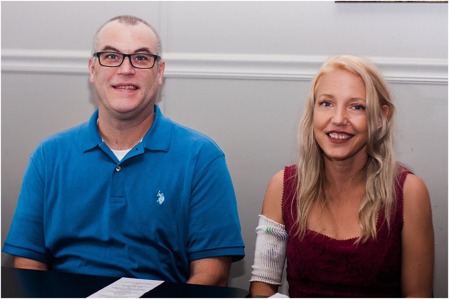Two guests sit together at an event, one wearing a blue polo and another in a burgundy dress.