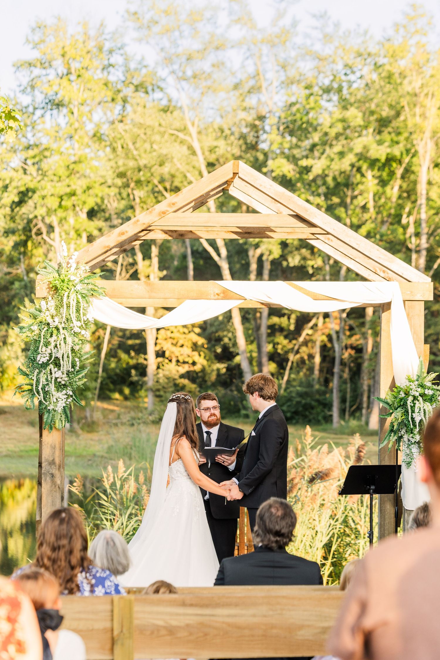Outdoor wedding ceremony under wooden pergola decorated with greenery during golden hour.