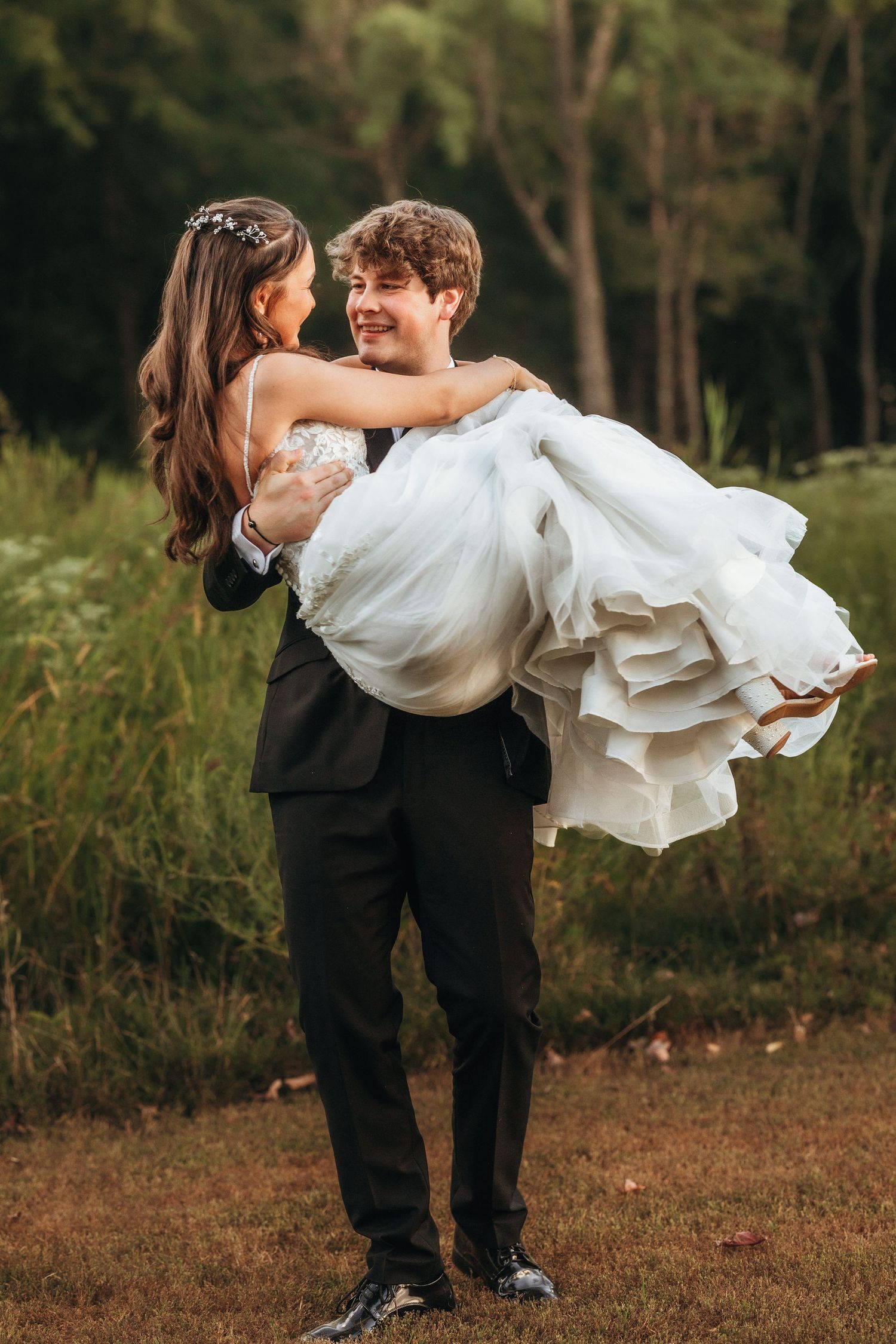 A romantic wedding moment captured as a groom carries his bride in a flowing white dress in a wooded outdoor setting.