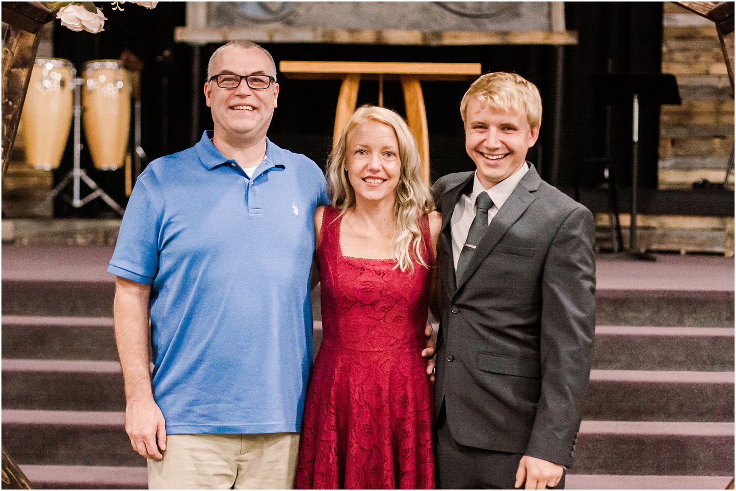 Three people in contrasting casual and formal attire pose at a rustic venue entrance.