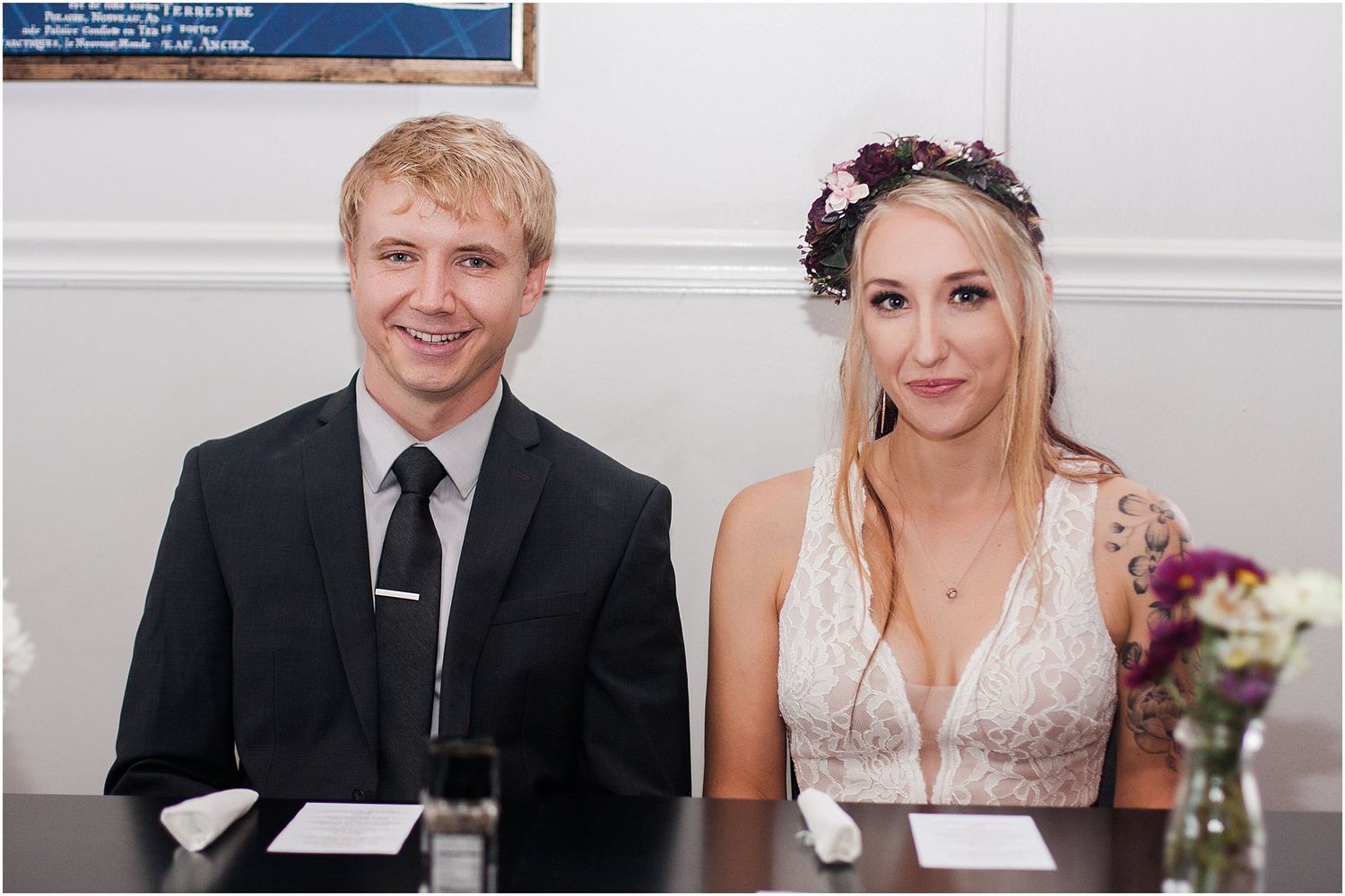 A couple sits at a table during what appears to be a wedding signing ceremony with flowers in the background.