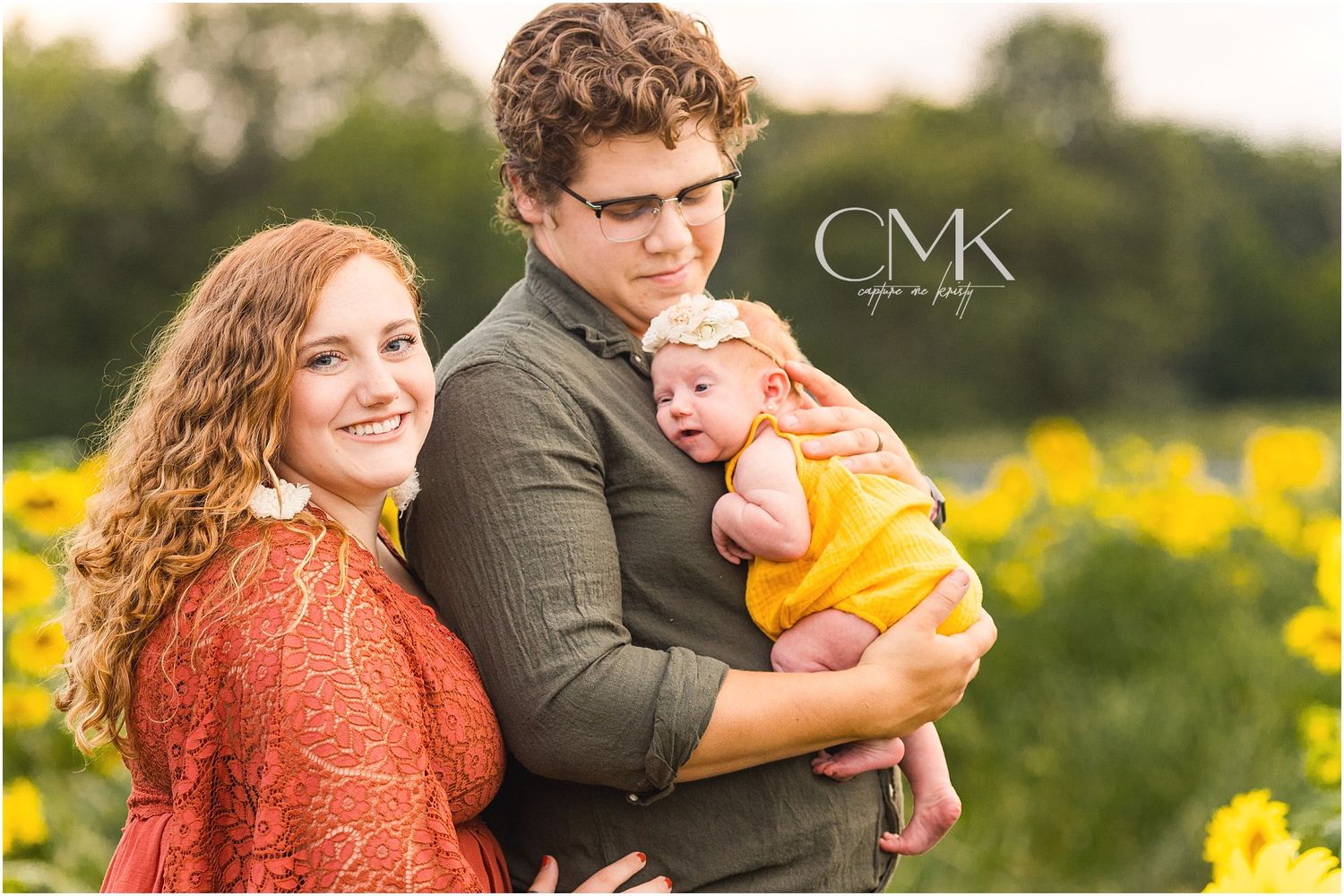 A couple holds their baby in a yellow outfit against a backdrop of yellow wildflowers at sunset.