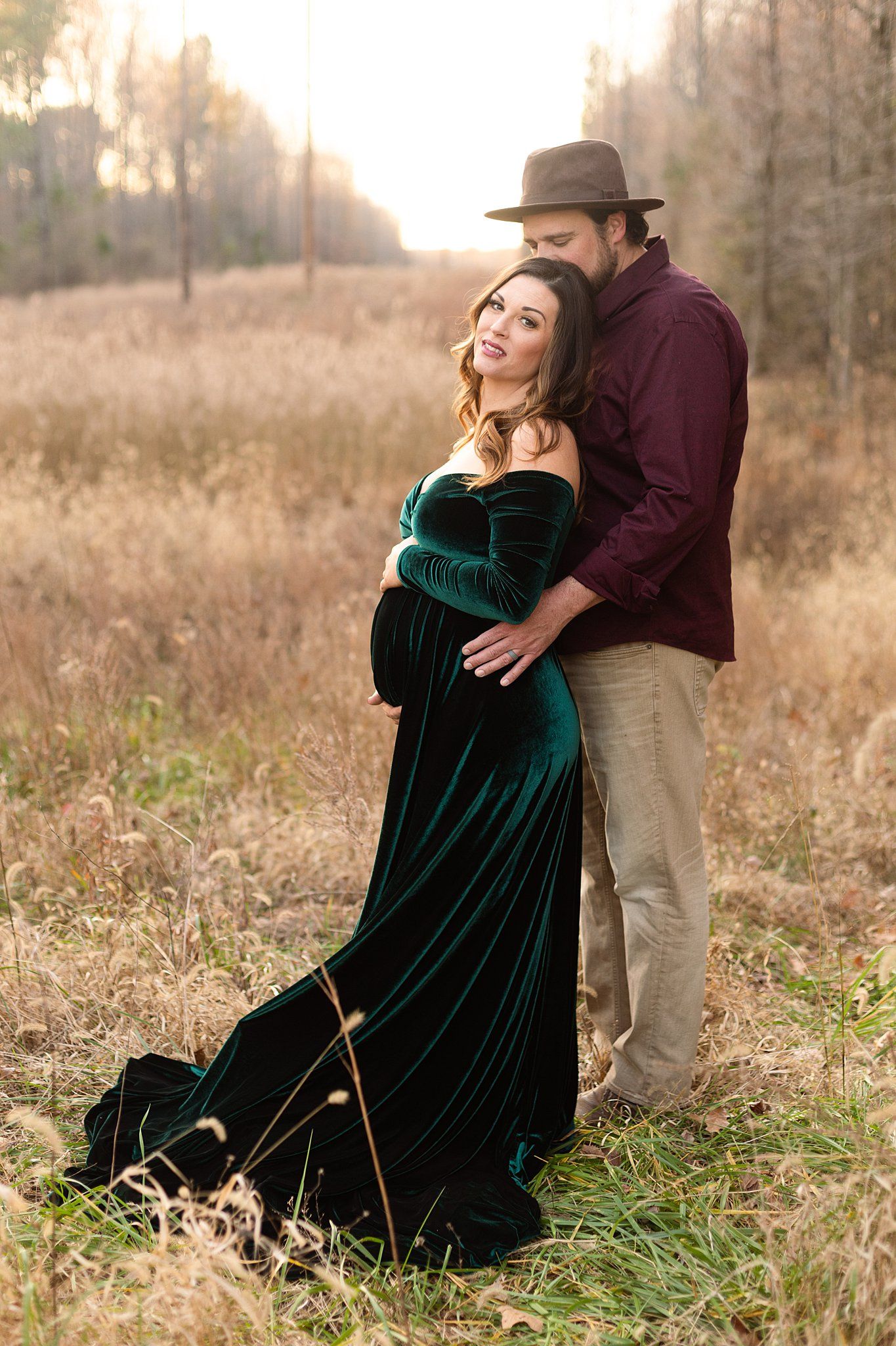 Romantic couple poses in autumn field with woman in flowing green velvet dress and man in burgundy shirt.