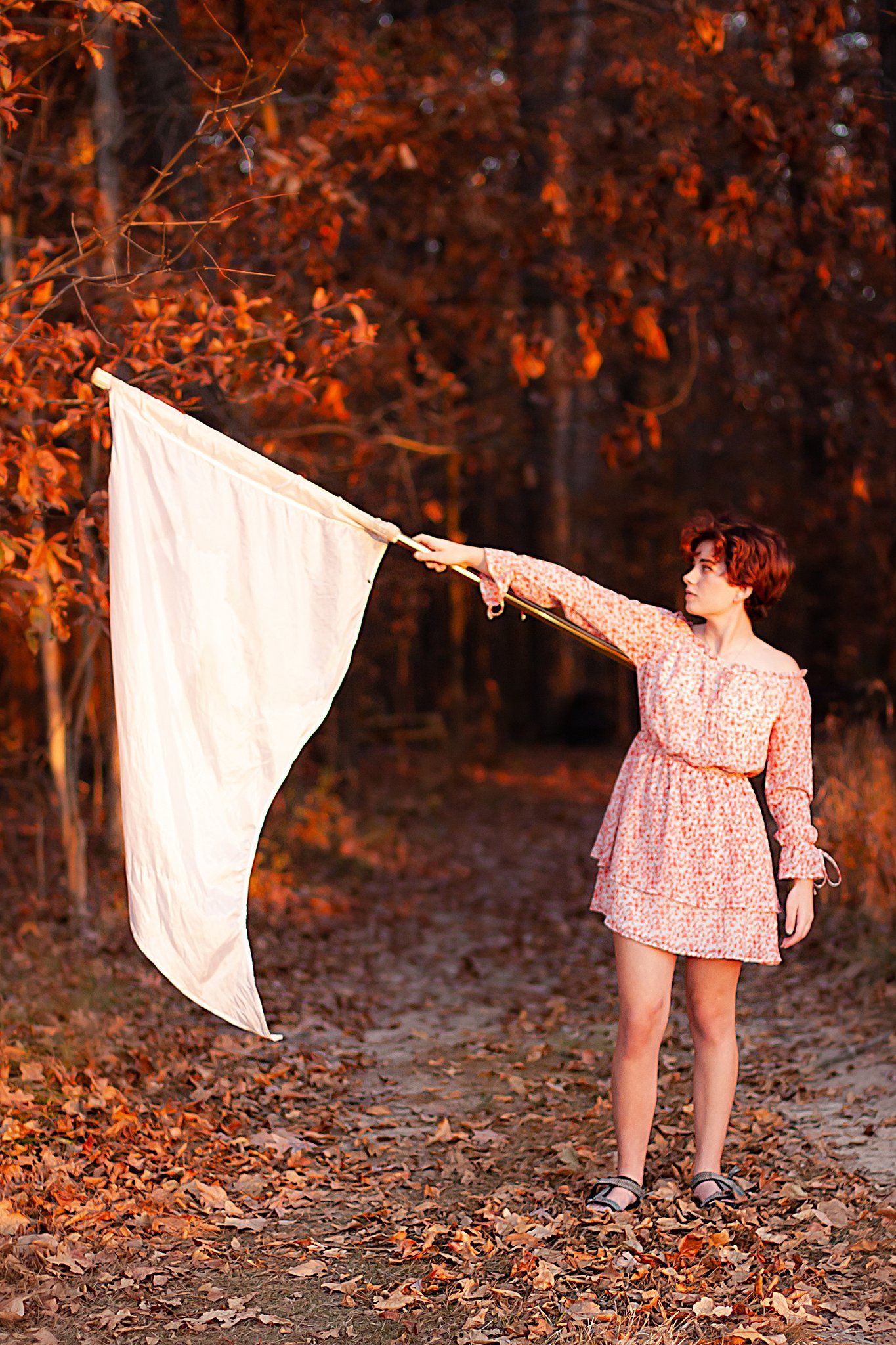 Person in floral dress holds white flag in autumn forest with orange foliage at sunset.
