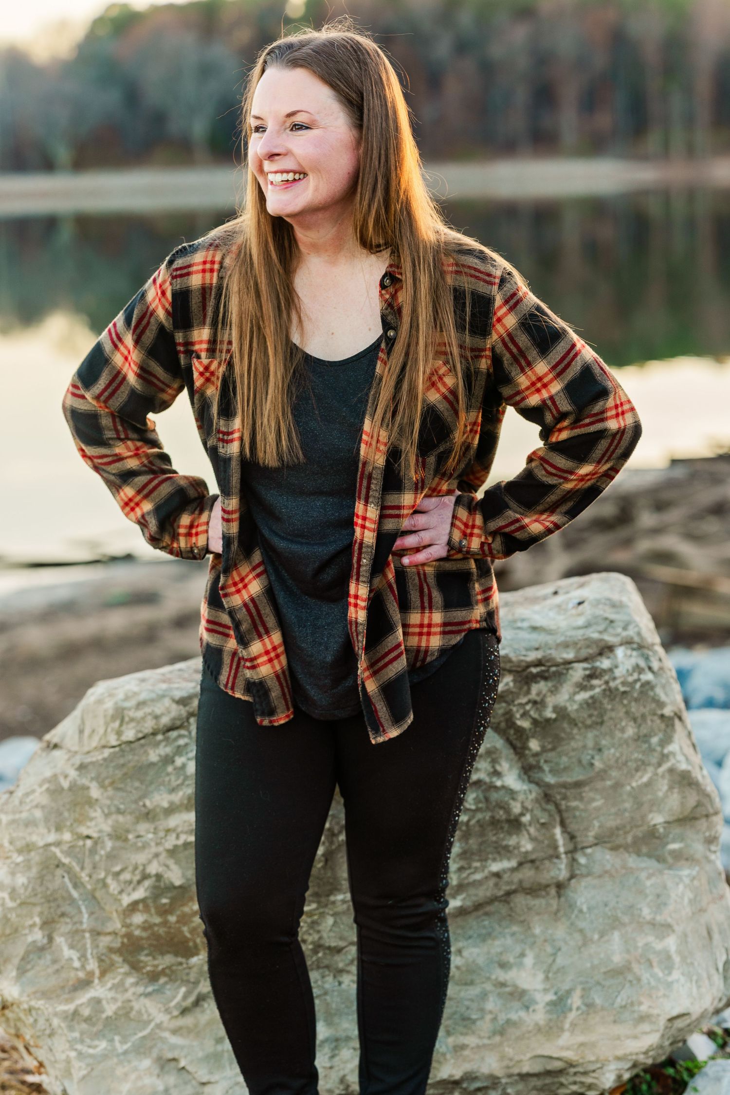Person in plaid flannel and black outfit poses on large rocks near water.