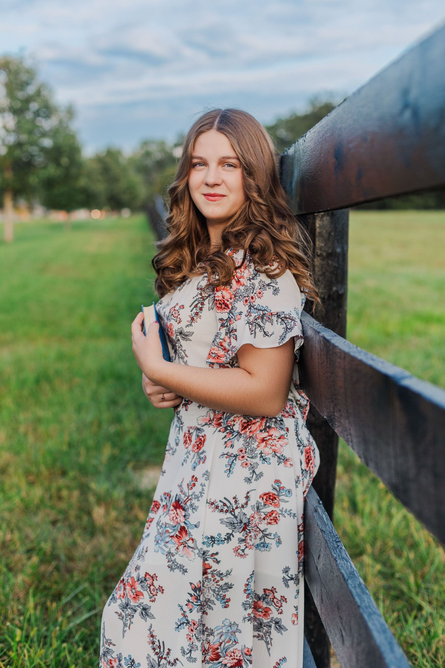 A country-style portrait in a floral dress leaning against a wooden fence.