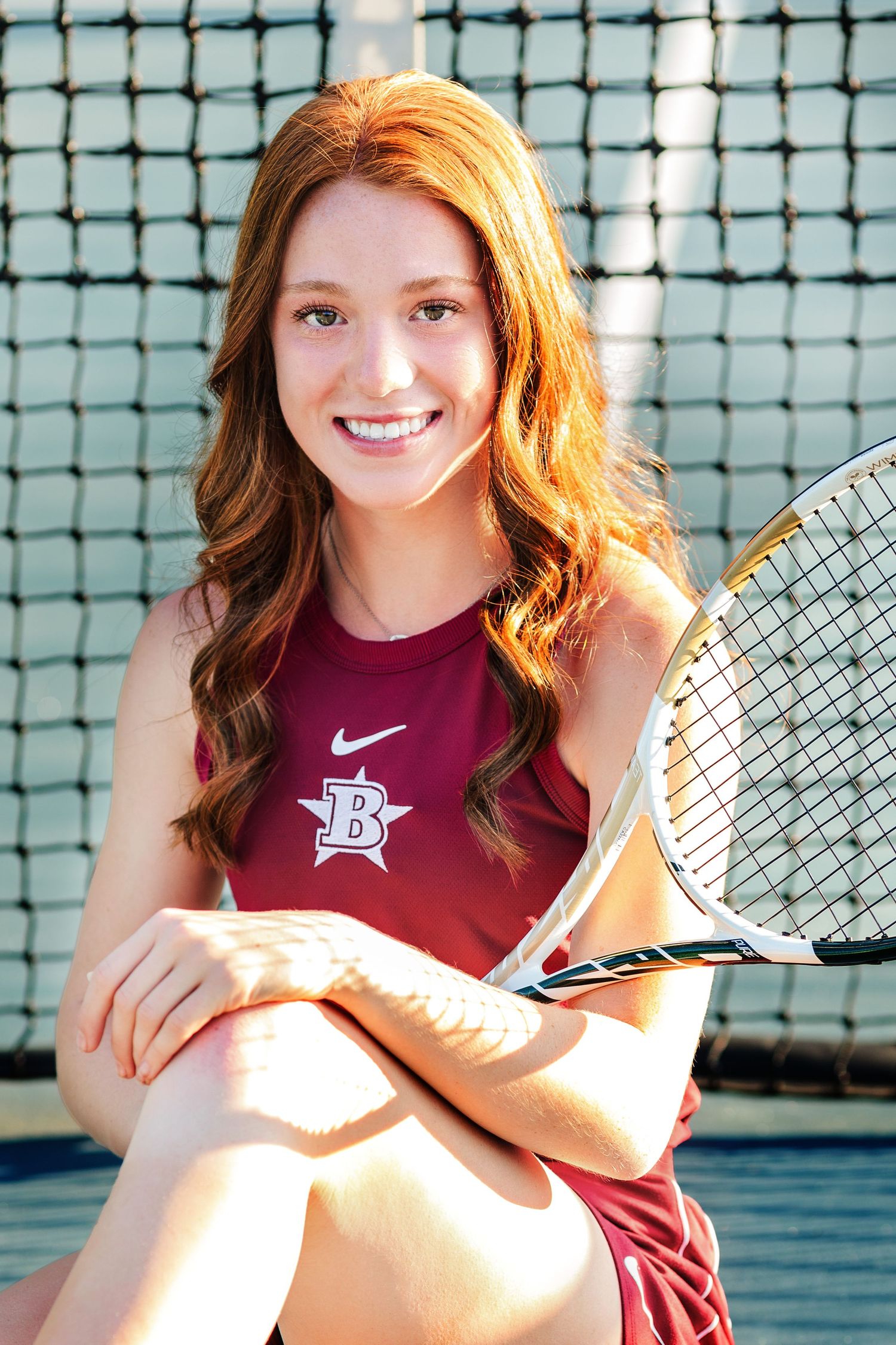Tennis player in maroon athletic wear poses with racquet against court netting.