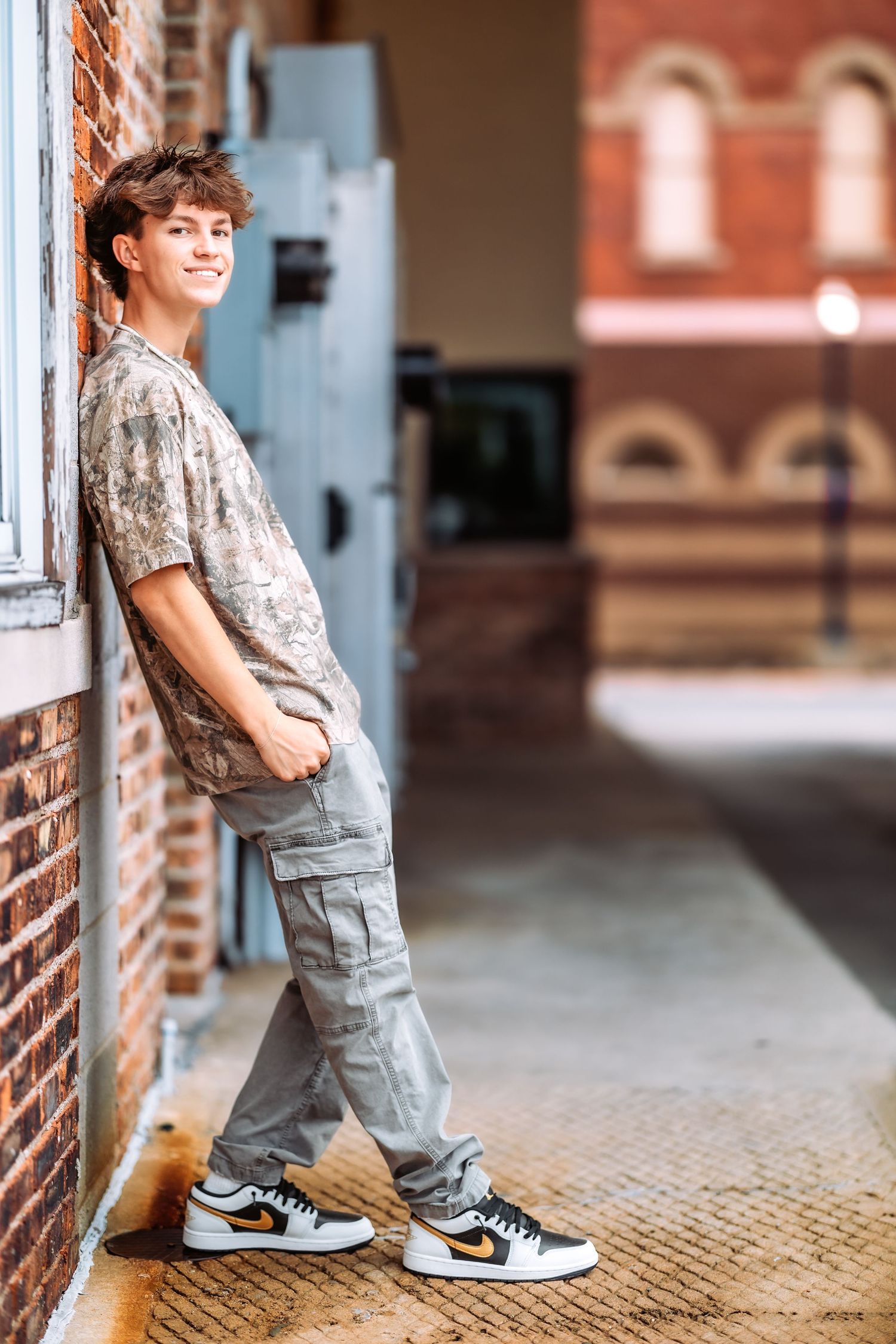 Child in patterned shirt and cargo pants poses near brick wall wearing sneakers.