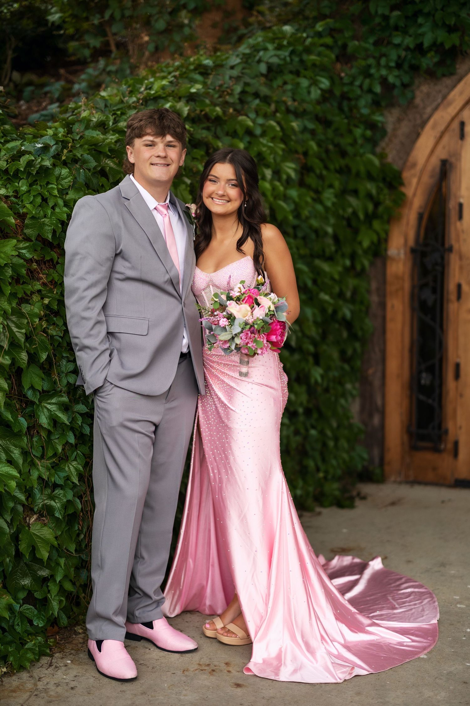 A couple poses in coordinating pink prom attire, the girl in a satin gown holding a floral bouquet outdoors.