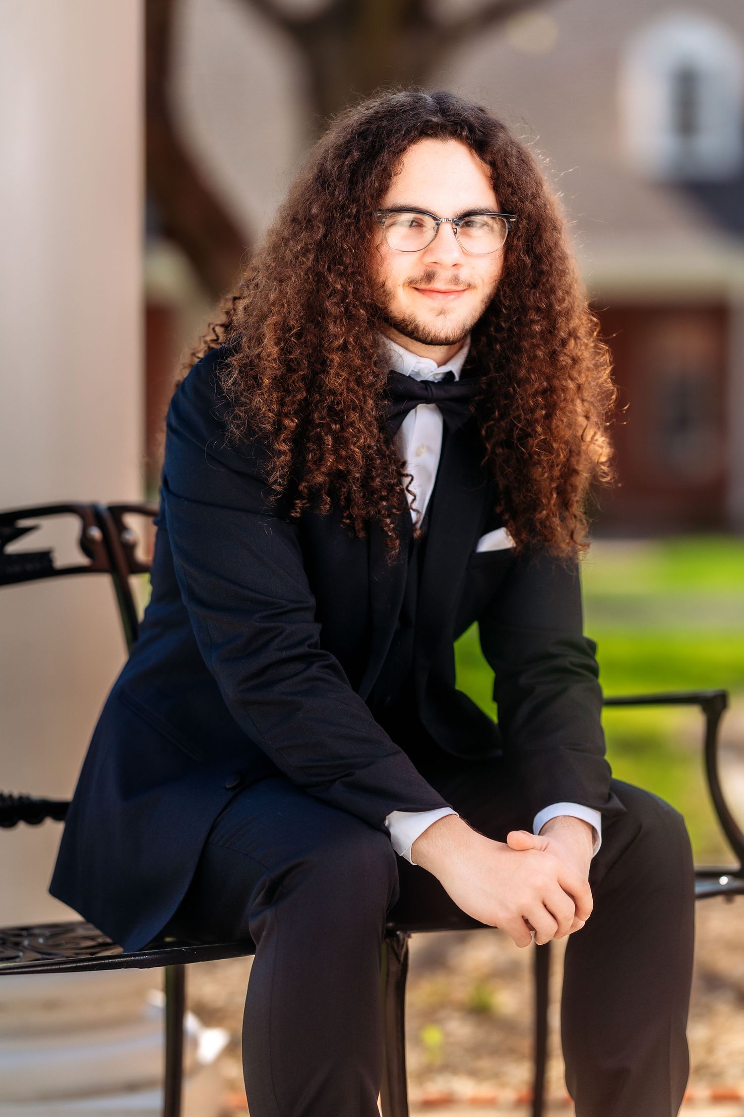 A graduate in a black suit and bow tie sits on a bench outdoors for a series of formal portraits.