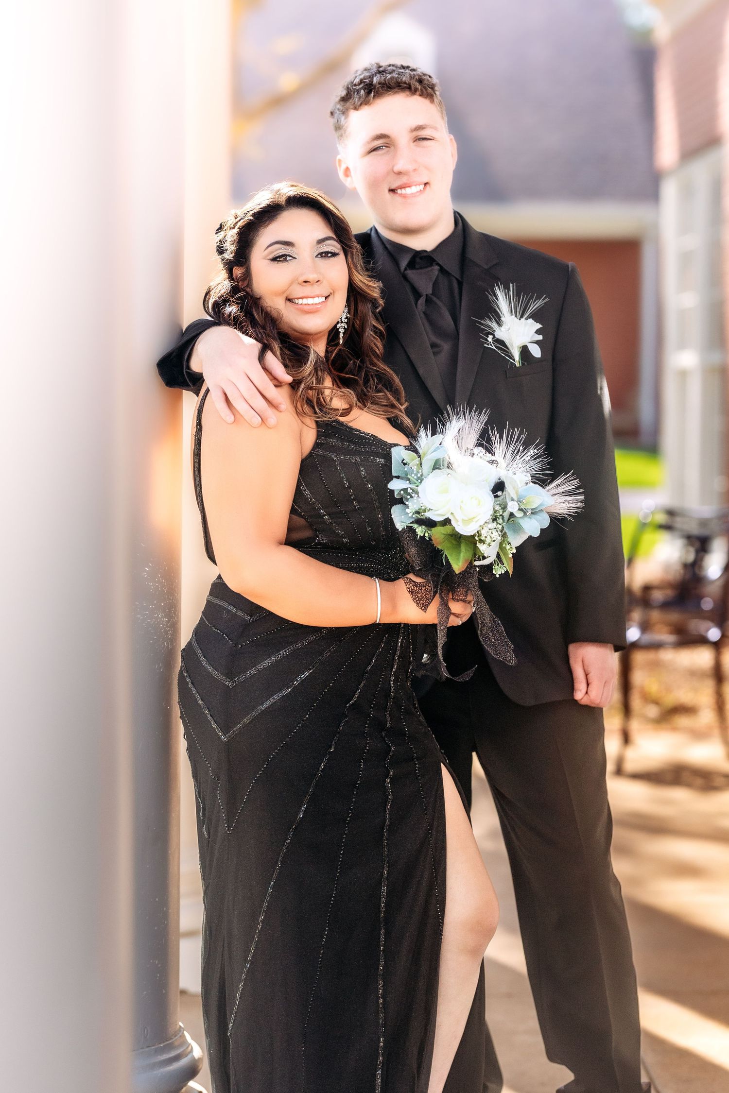 A couple in formal black attire pose together at a wedding with white floral boutonnieres and bouquets.