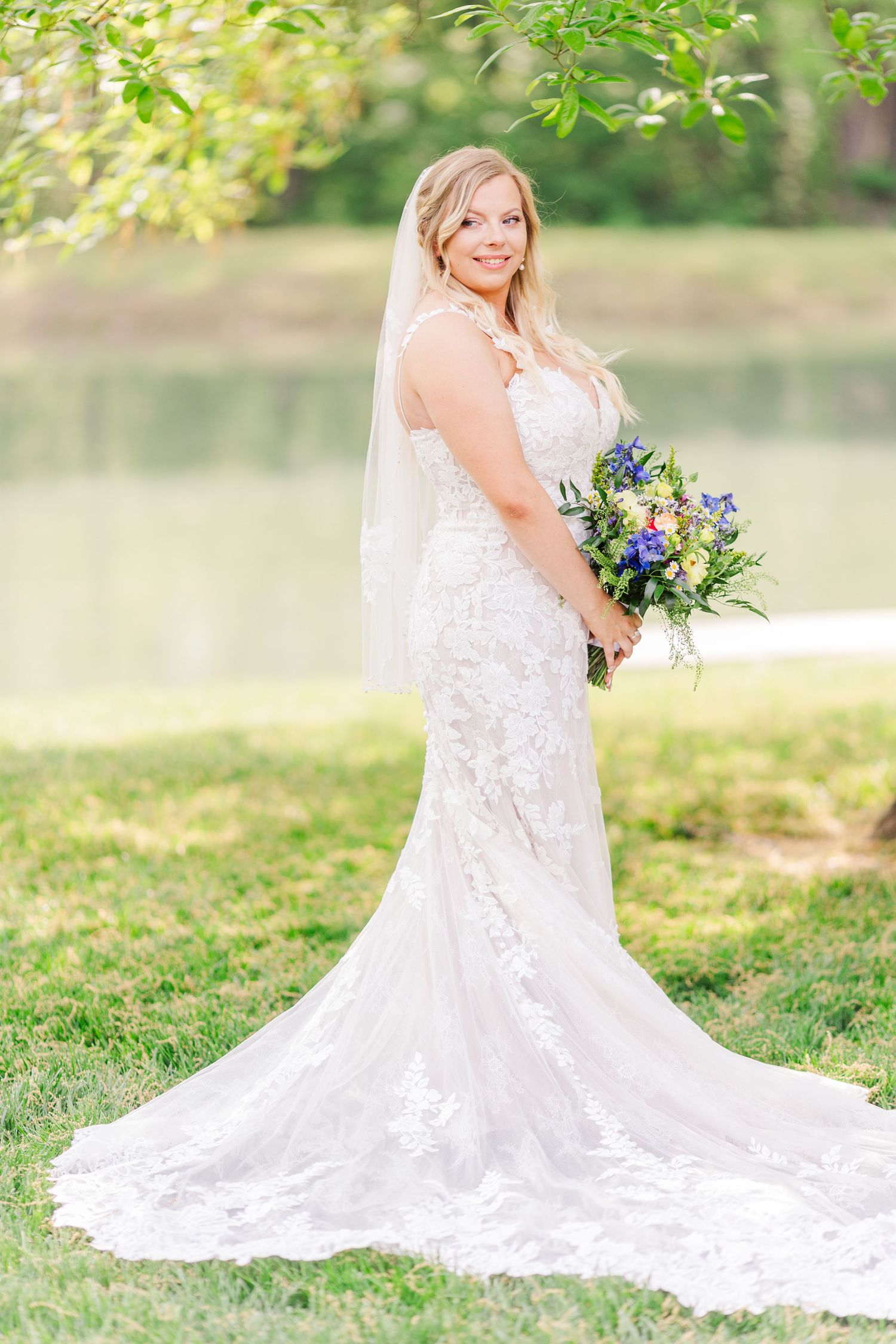 A bride in a flowing lace wedding dress poses outdoors with a vibrant blue and purple bouquet.