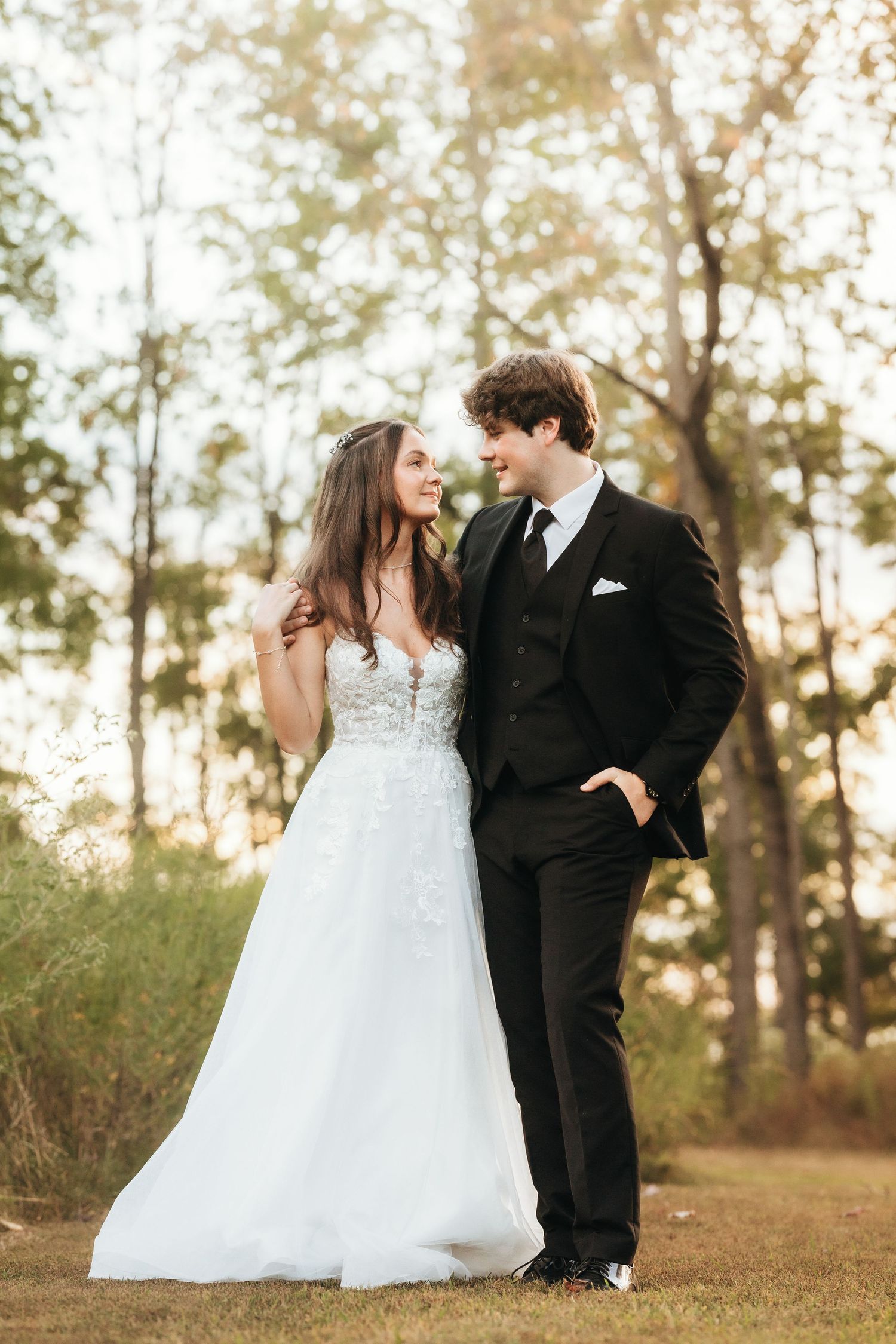 Elegant wedding portrait of couple in formal attire against a sunlit forest backdrop.