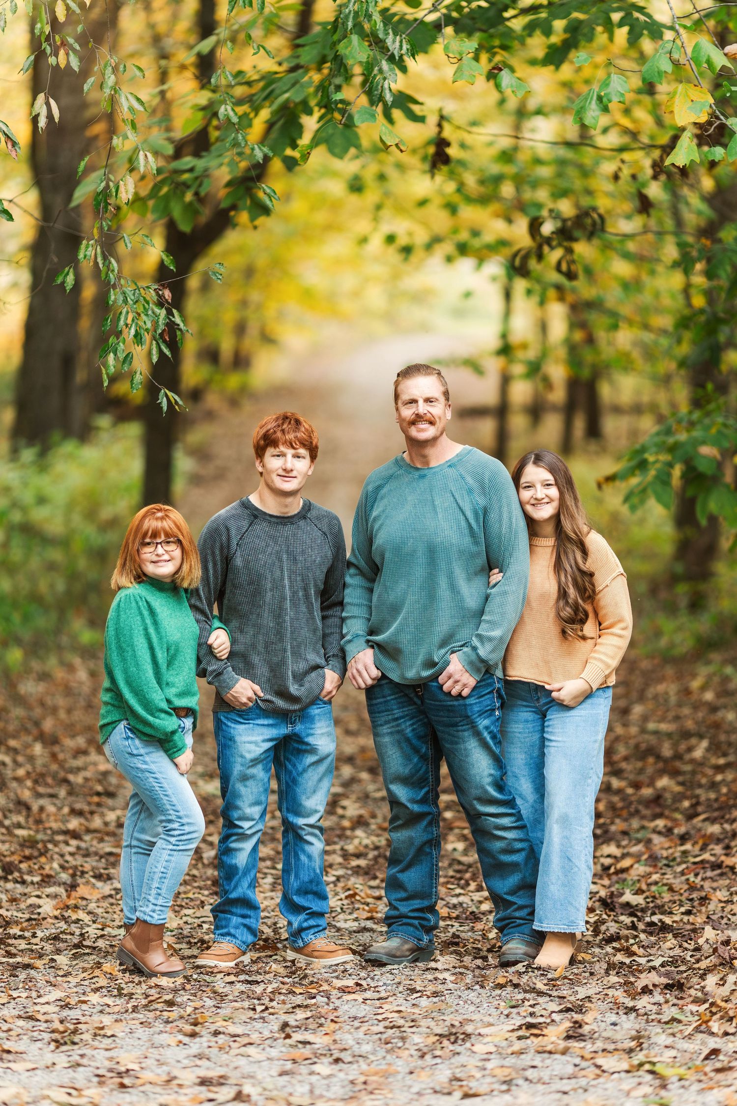 A casual family portrait shows five people in coordinated sweaters standing on an autumn forest path.