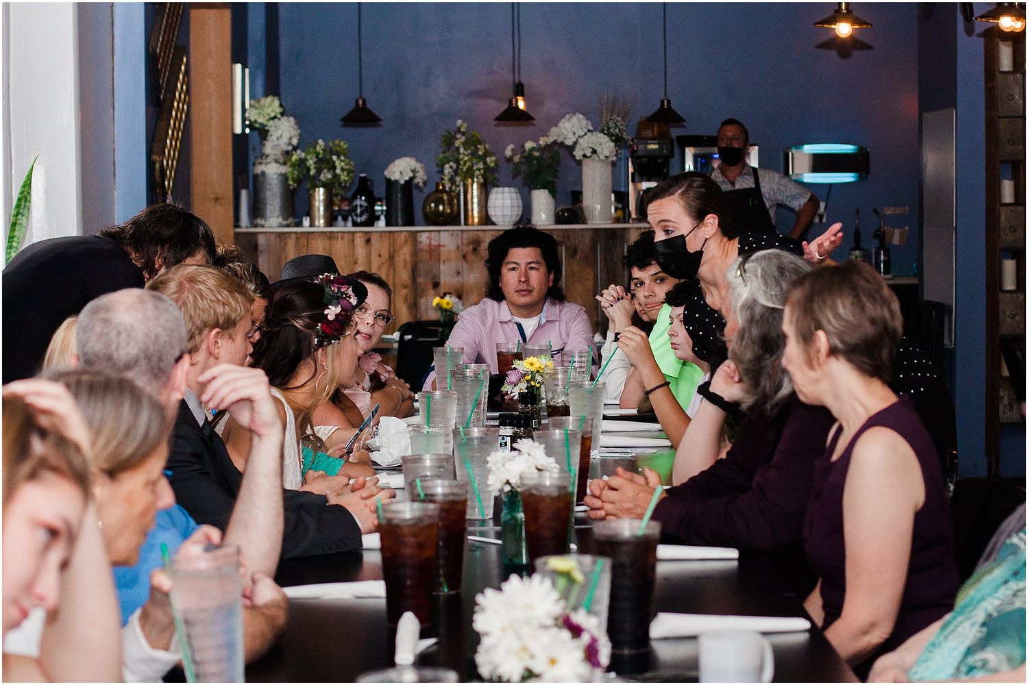 A group of wedding guests enjoying dinner and conversation at a long table in a restaurant setting.