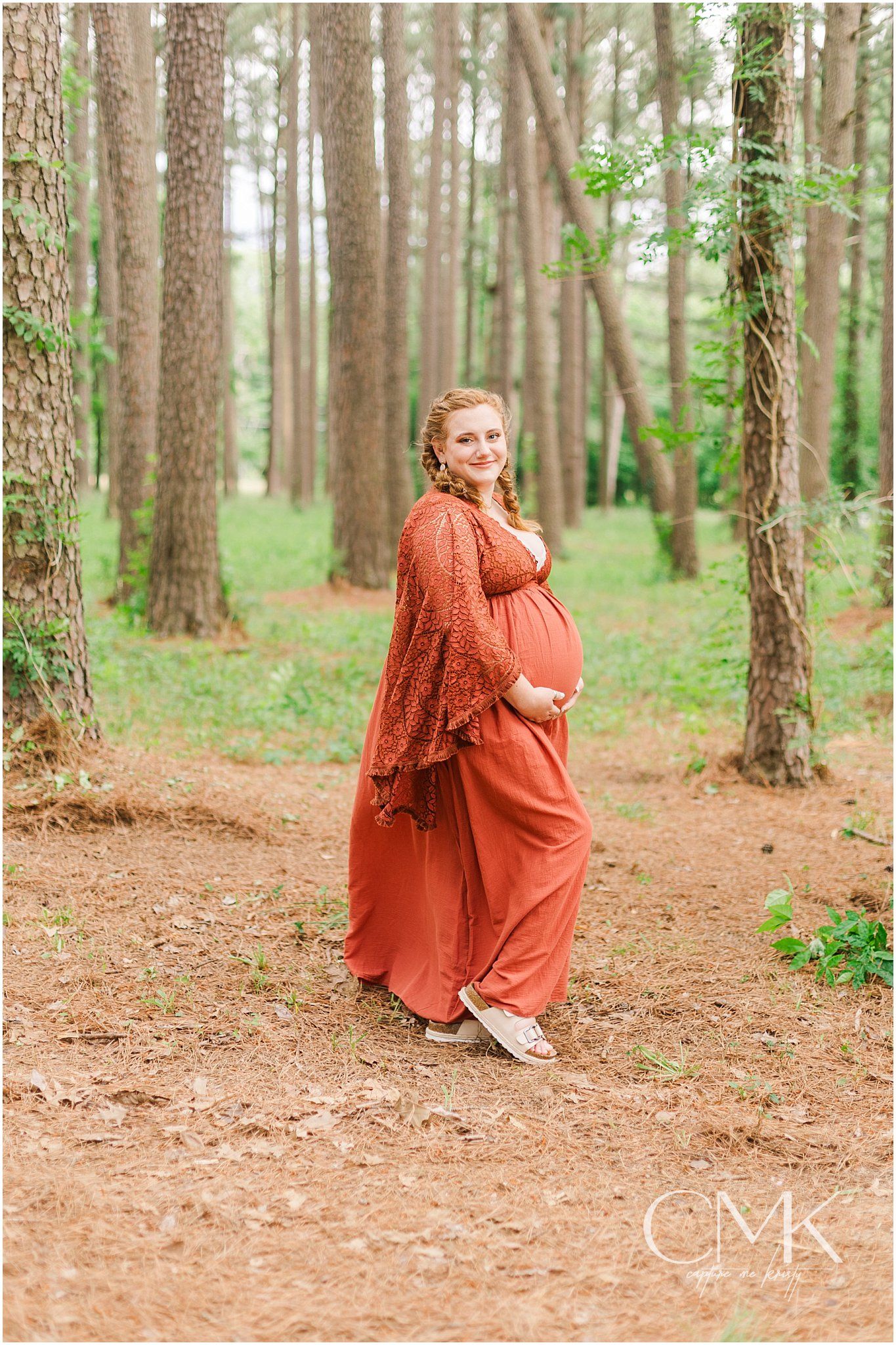 Full-length maternity photo in coral dress standing among tall pine trees.