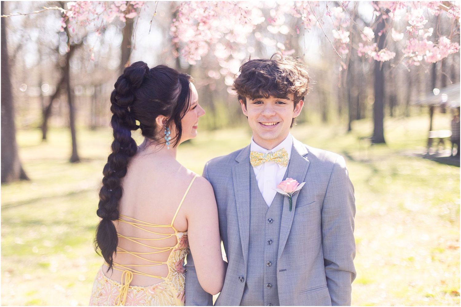 Couple in prom attire posing in spring garden with blooming cherry trees.