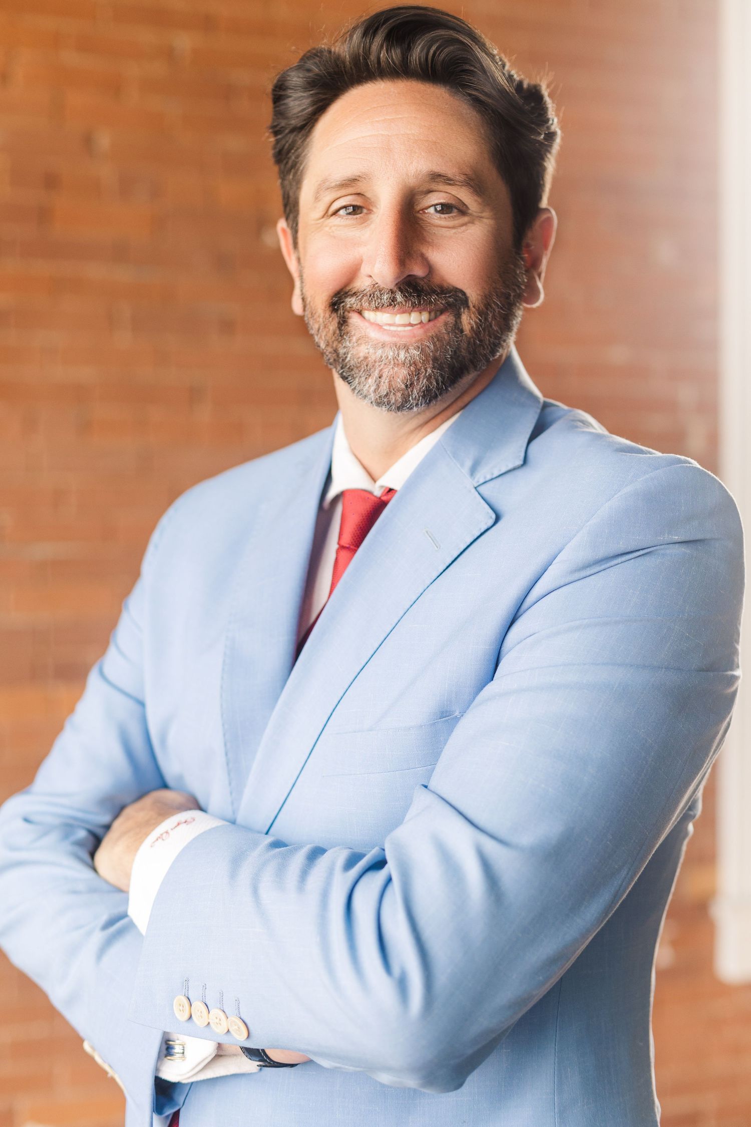 Professional portrait of person wearing light blue blazer and red tie against brick wall.