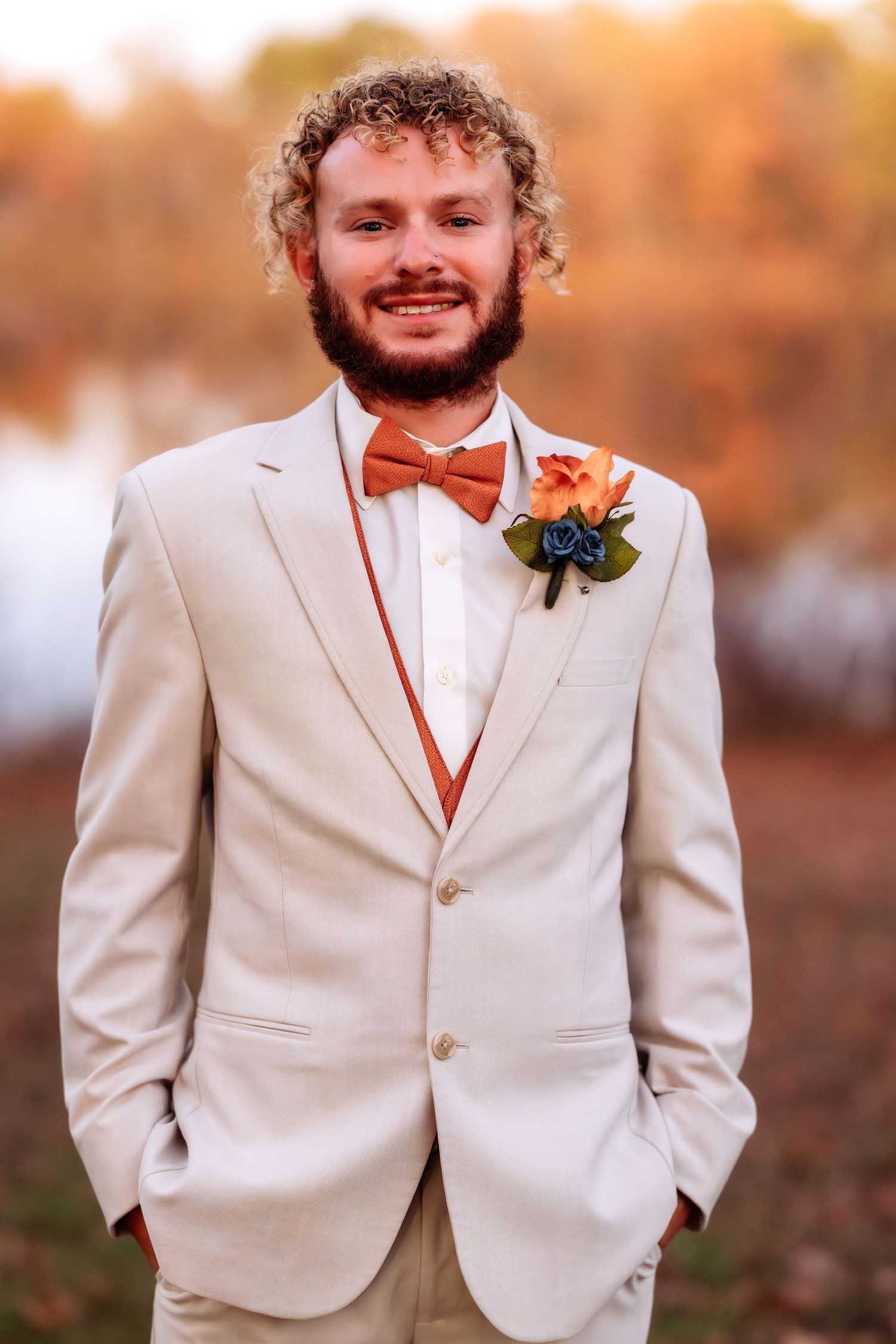 Groom in a cream-colored suit with boutonniere smiling during outdoor autumn wedding.