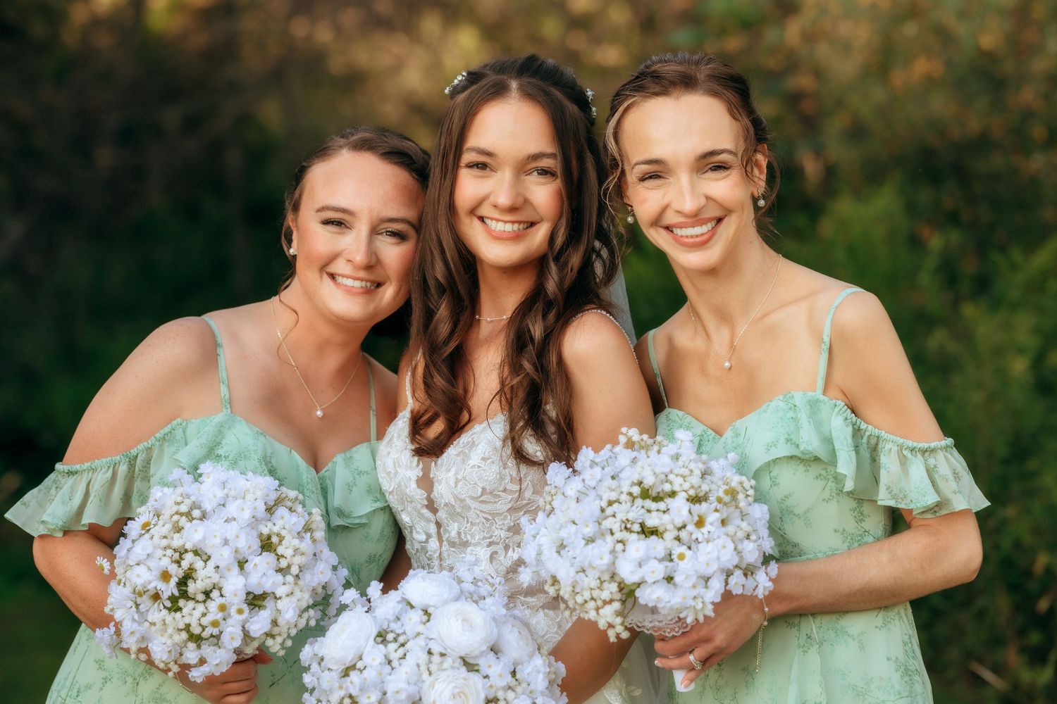 Two bridesmaids in mint green dresses pose together with the bride, holding white floral bouquets outdoors.
