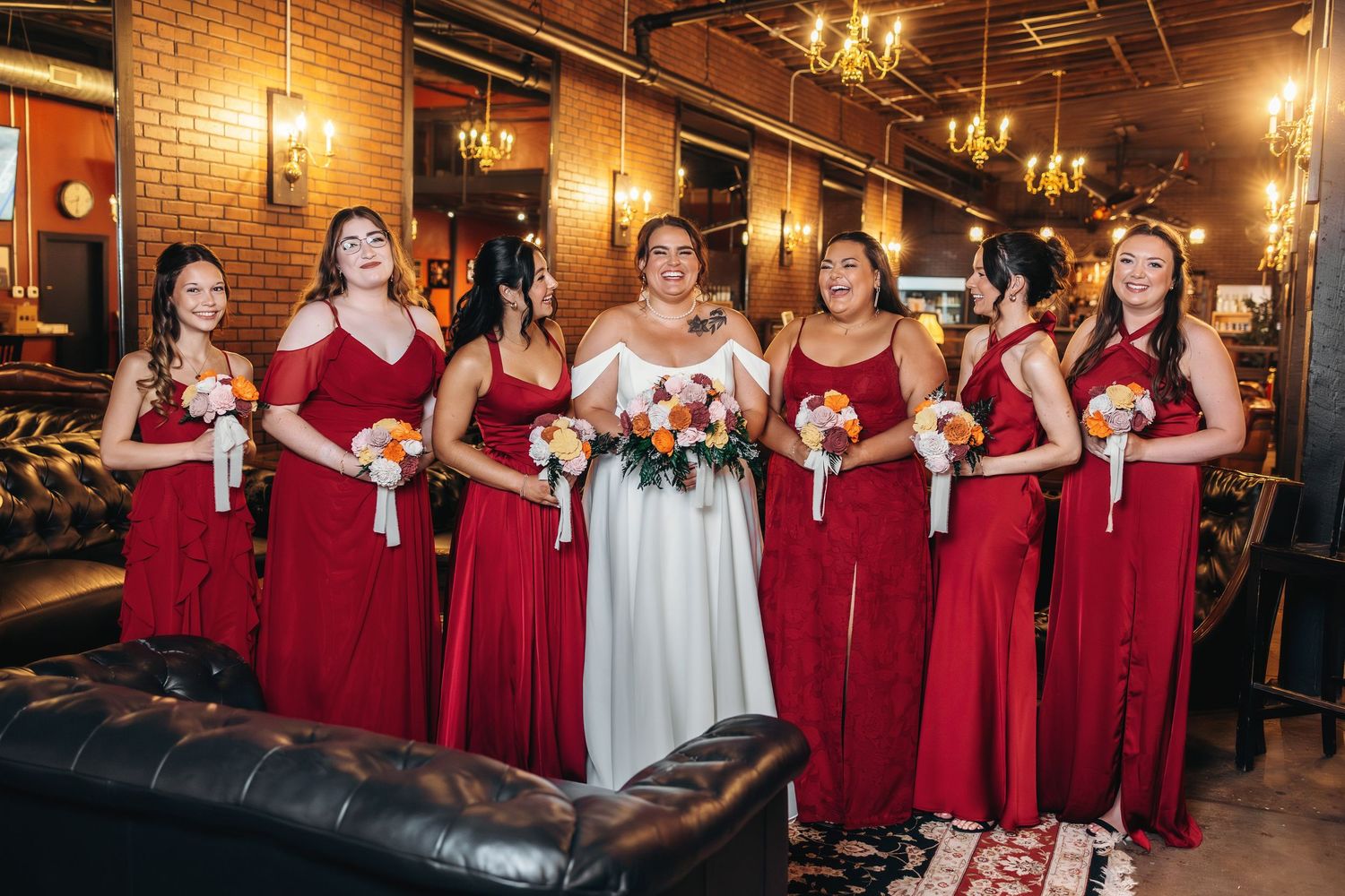 Bridesmaids in burgundy dresses gather in an industrial-style venue with brick walls.