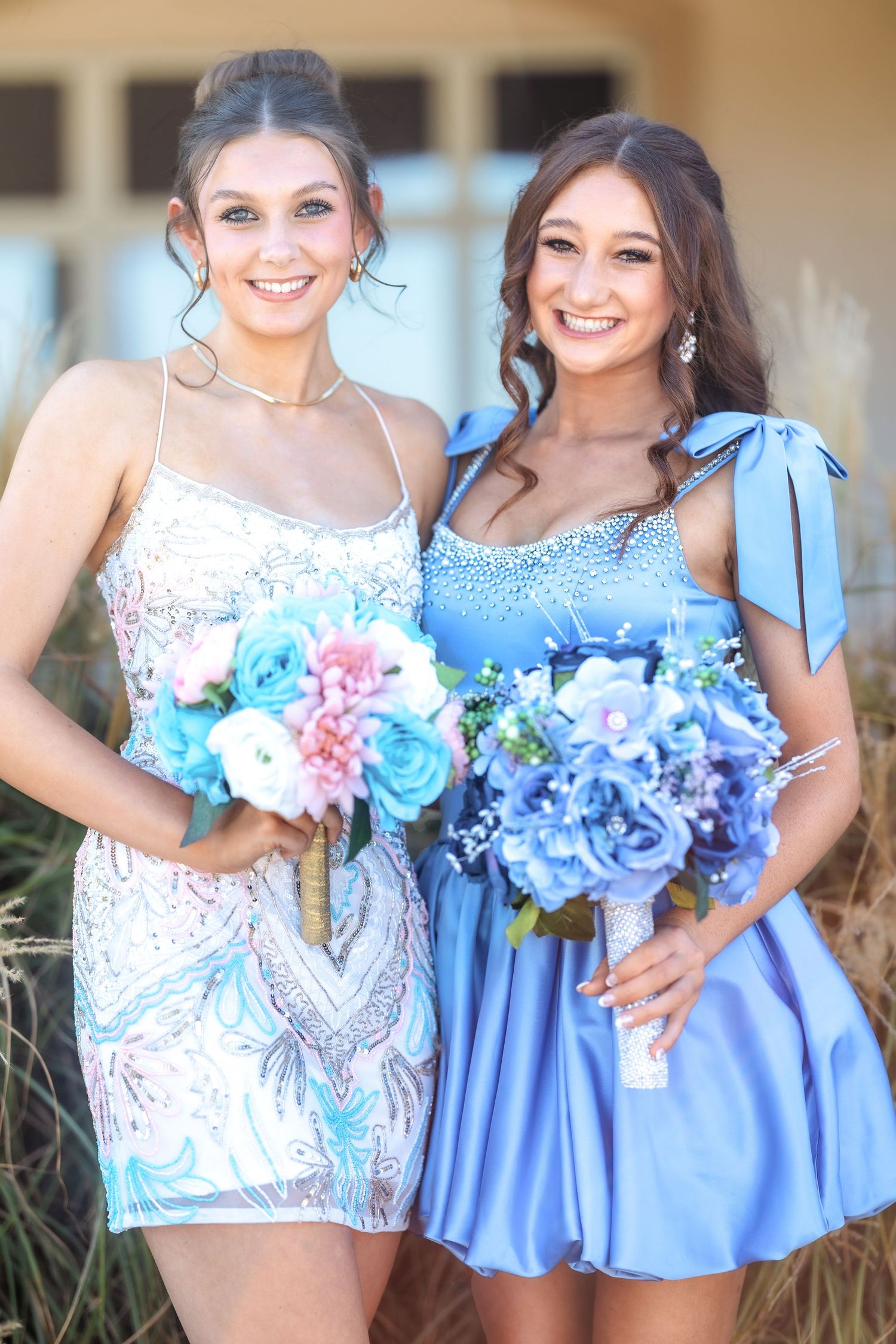 Two women in formal dresses hold blue and pink floral bouquets while posing together.
