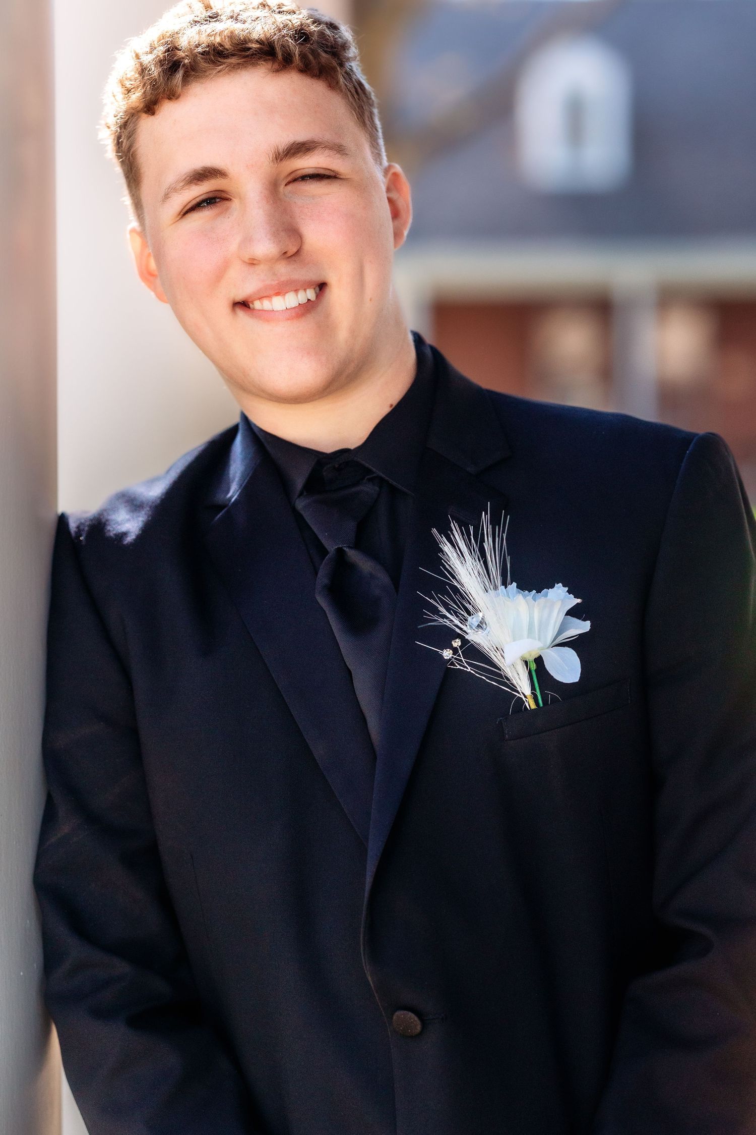 A person in a black suit jacket and tie smiles warmly while wearing a light blue and white feather boutonniere.