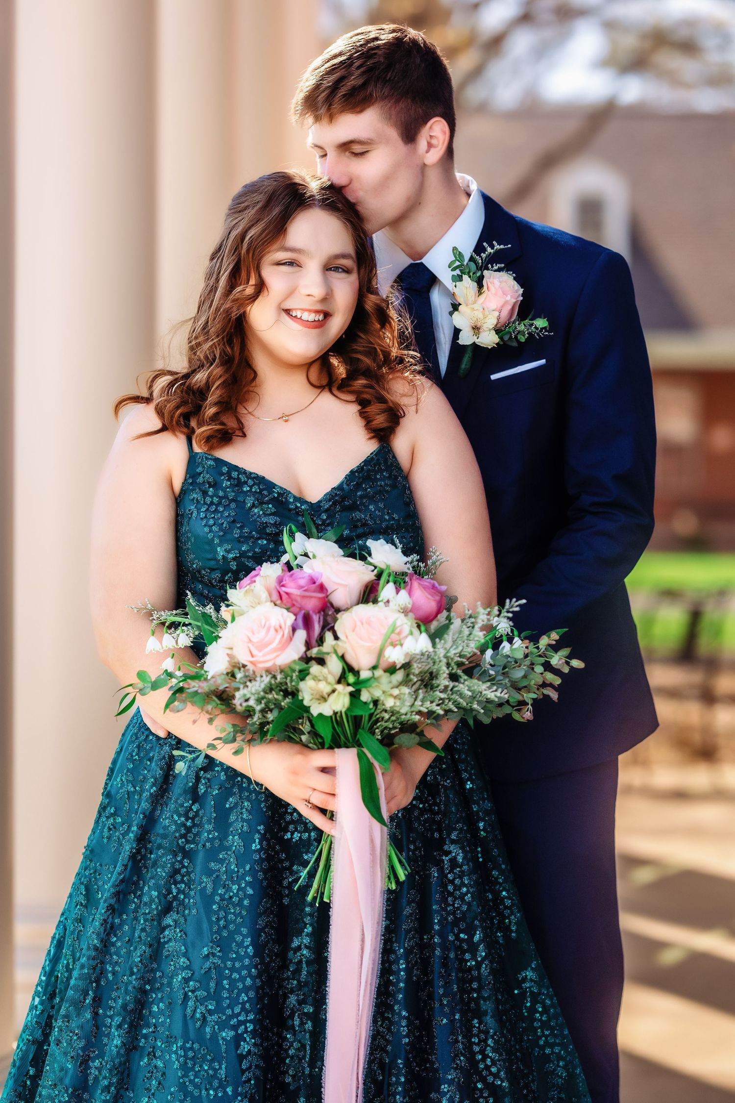 Wedding couple poses together in formal attire with emerald green dress and navy suit holding pink floral bouquet.