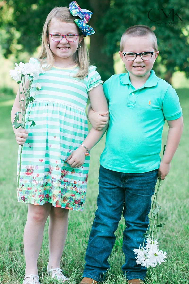 Two children in coordinating mint green outfits hold white flowers while standing on grass outdoors.