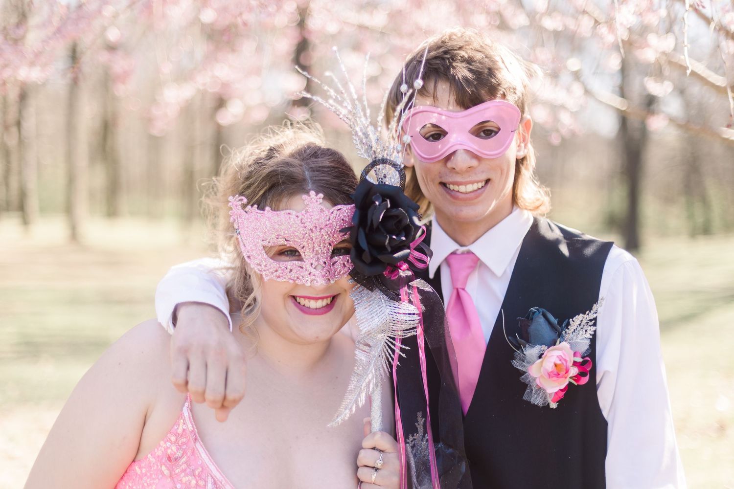 Couple wearing sparkly masks and formal attire pose under blooming spring trees.