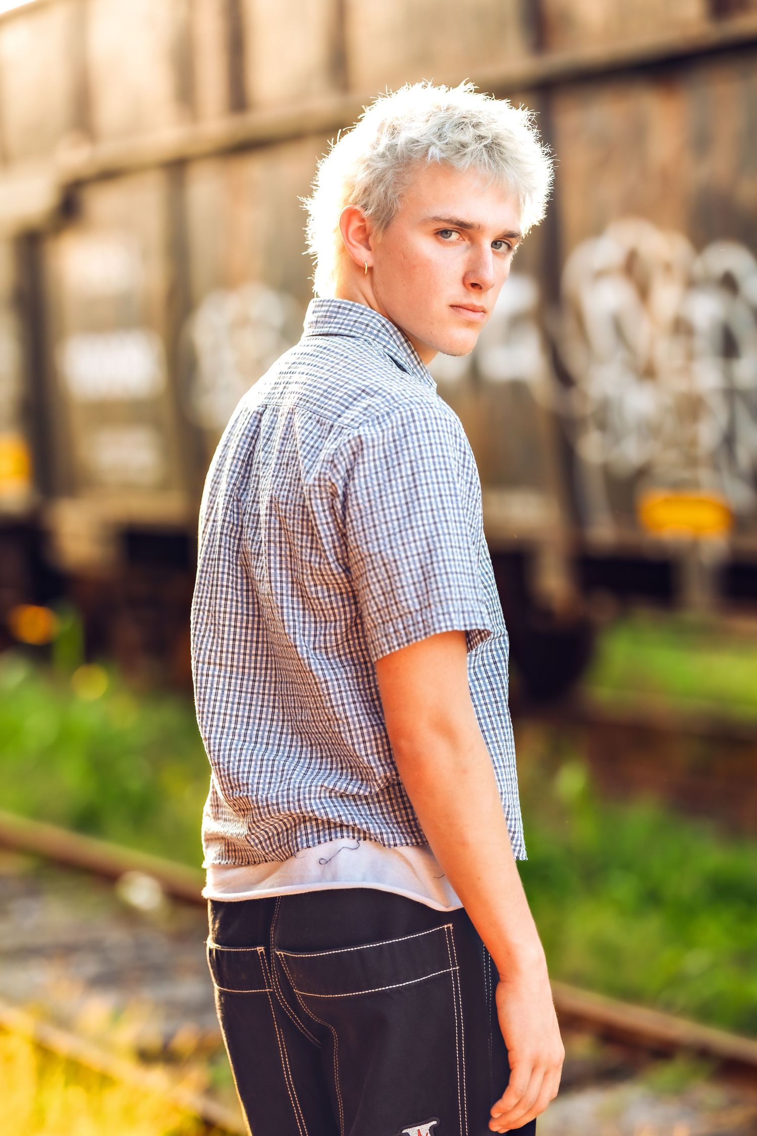 Individual in casual checkered shirt and jeans poses near industrial setting at sunset.
