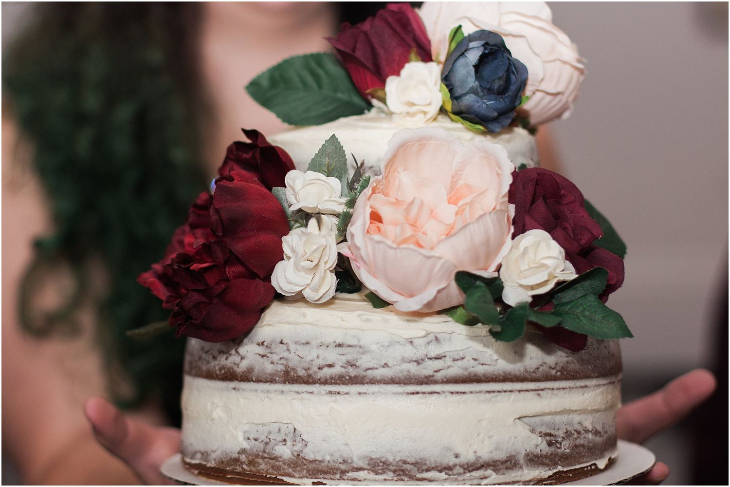 Close up of a semi-naked wedding cake with burgundy, blush and navy floral decorations.