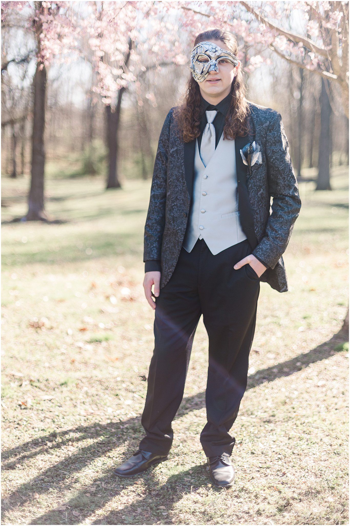 Person in dark suit with masquerade mask poses among spring trees.