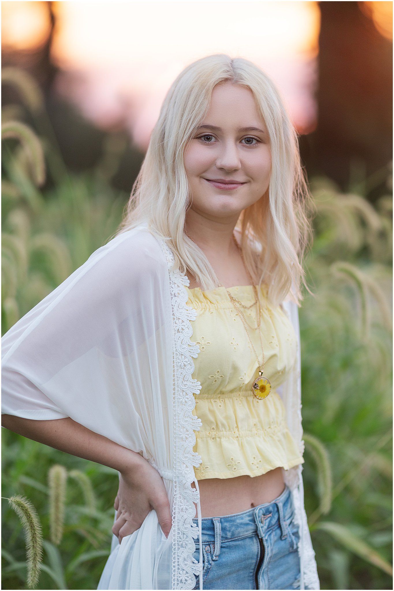 Senior girl with hands on hips with tall grass behind with sunset behind her