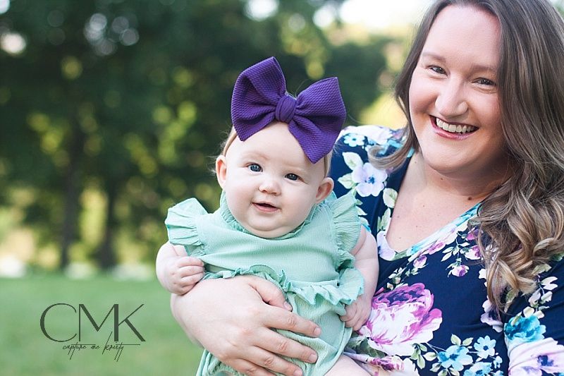 A floral-dressed woman holds a baby wearing mint green and a purple bow against a blurred green background.