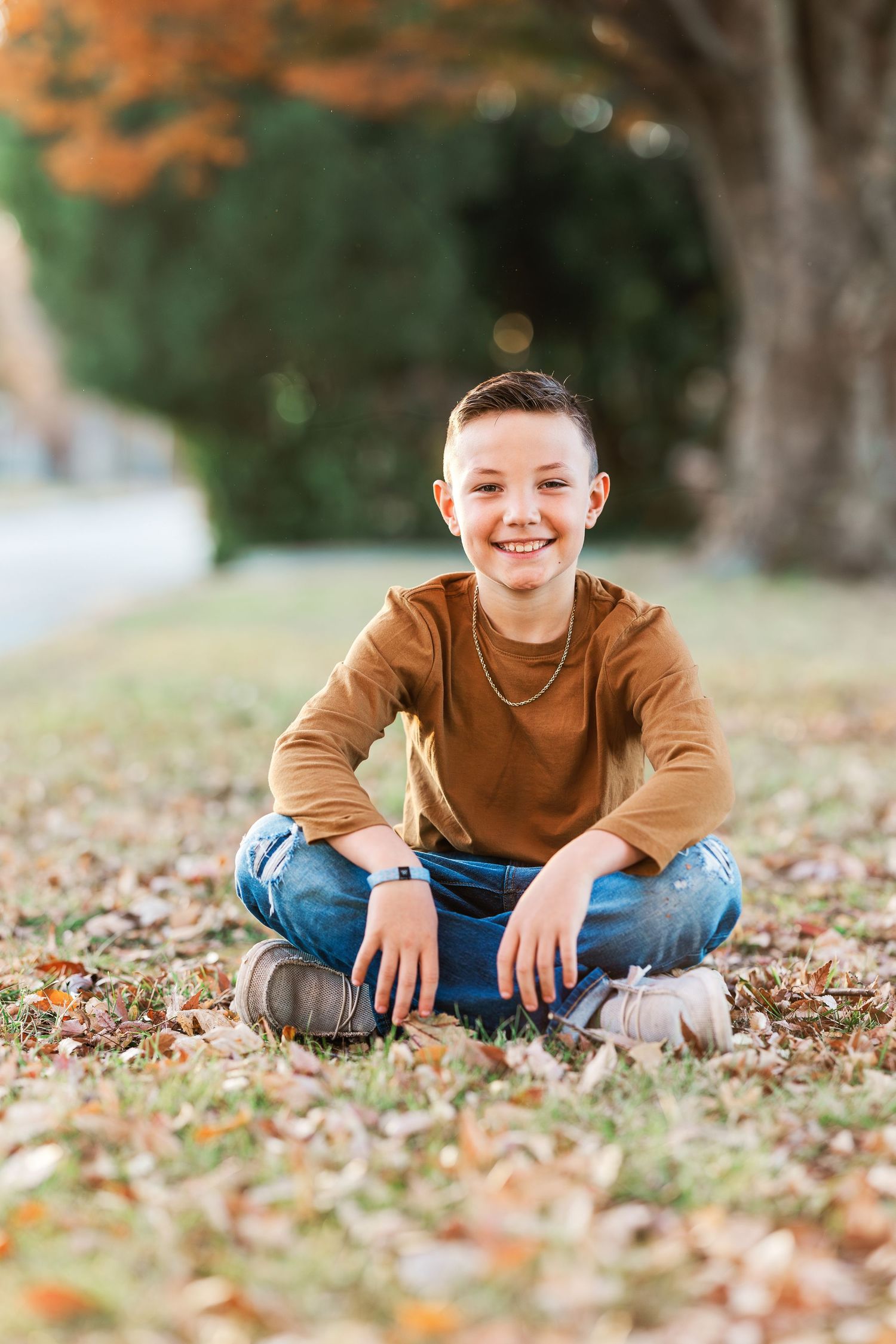 Child in brown sweater sits cross-legged on ground covered in autumn leaves.