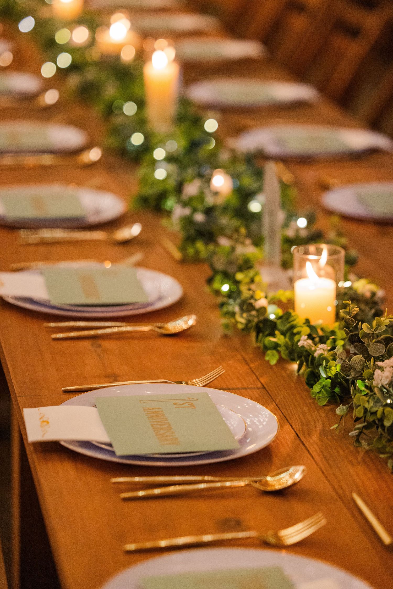 Elegant wedding reception table setting with greenery garland, candles and white plates on wooden table.