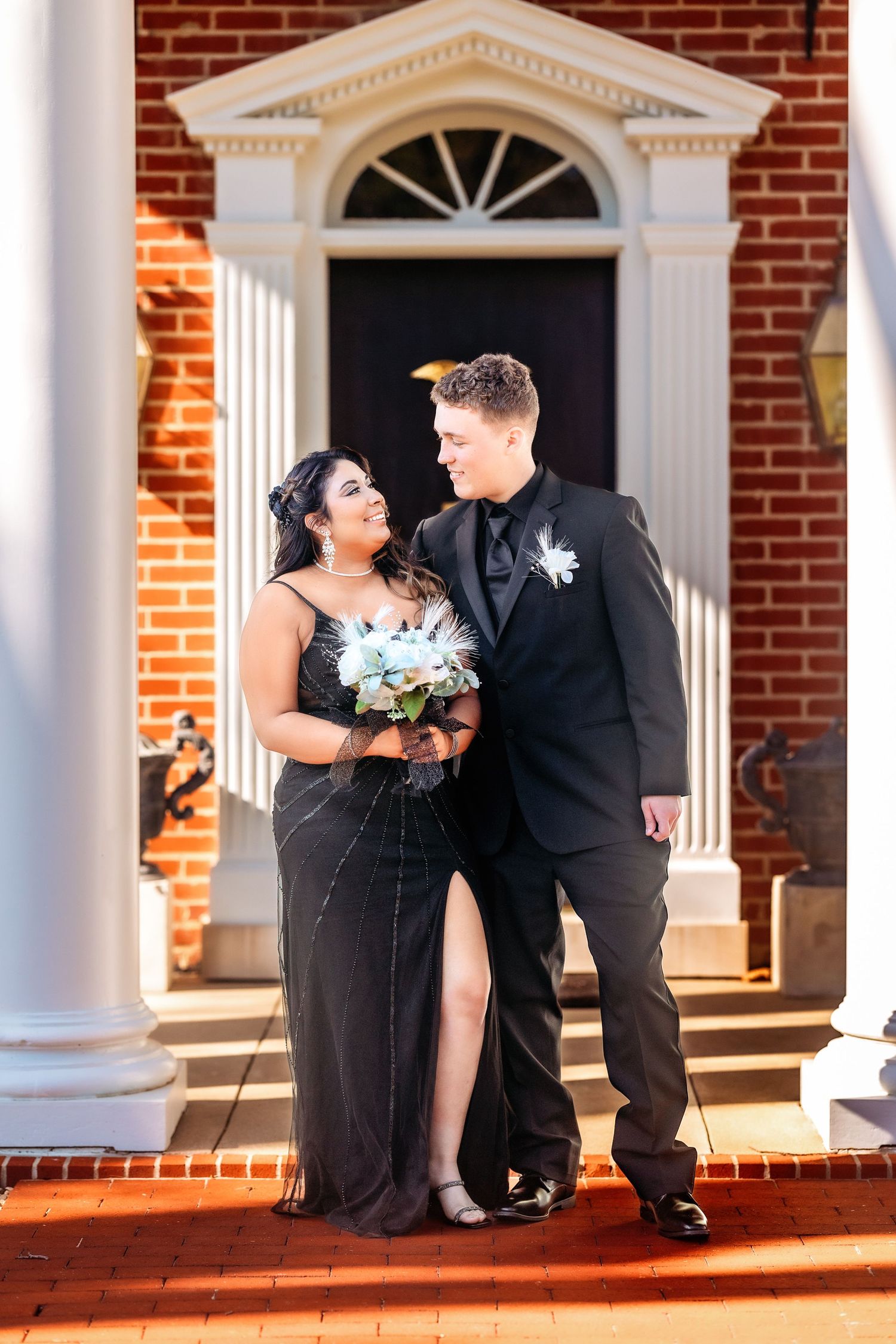 A couple in formal black attire shares a tender moment on the steps of a brick building with white columns.