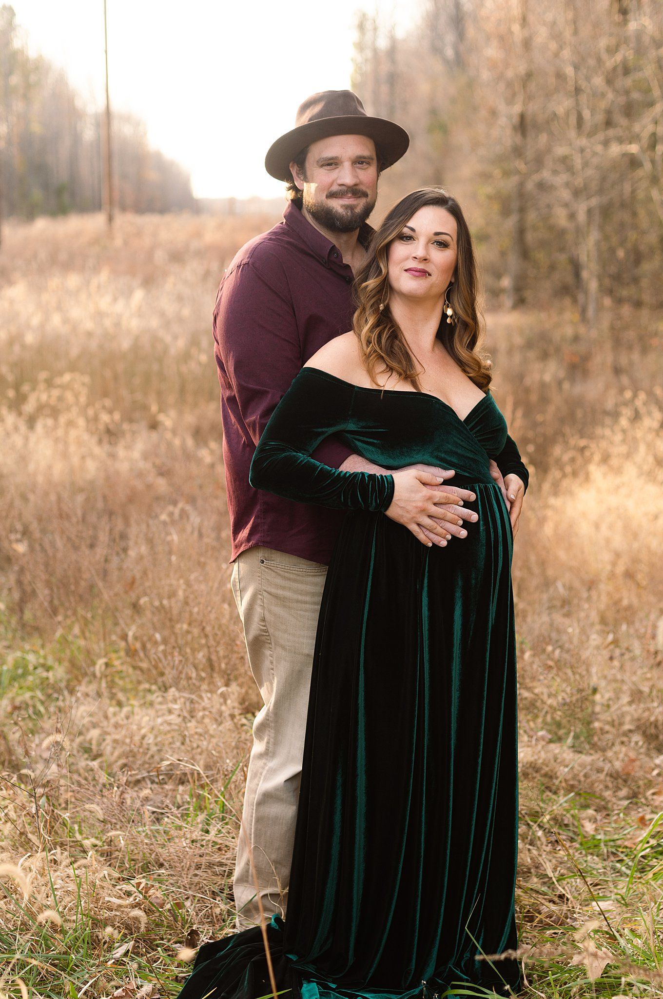 A maternity portrait in an autumn field featuring a green velvet dress and burgundy sweater.