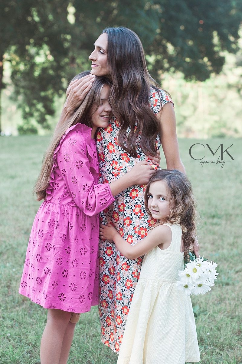Three people in floral and pink dresses share a loving embrace during an outdoor family photoshoot.