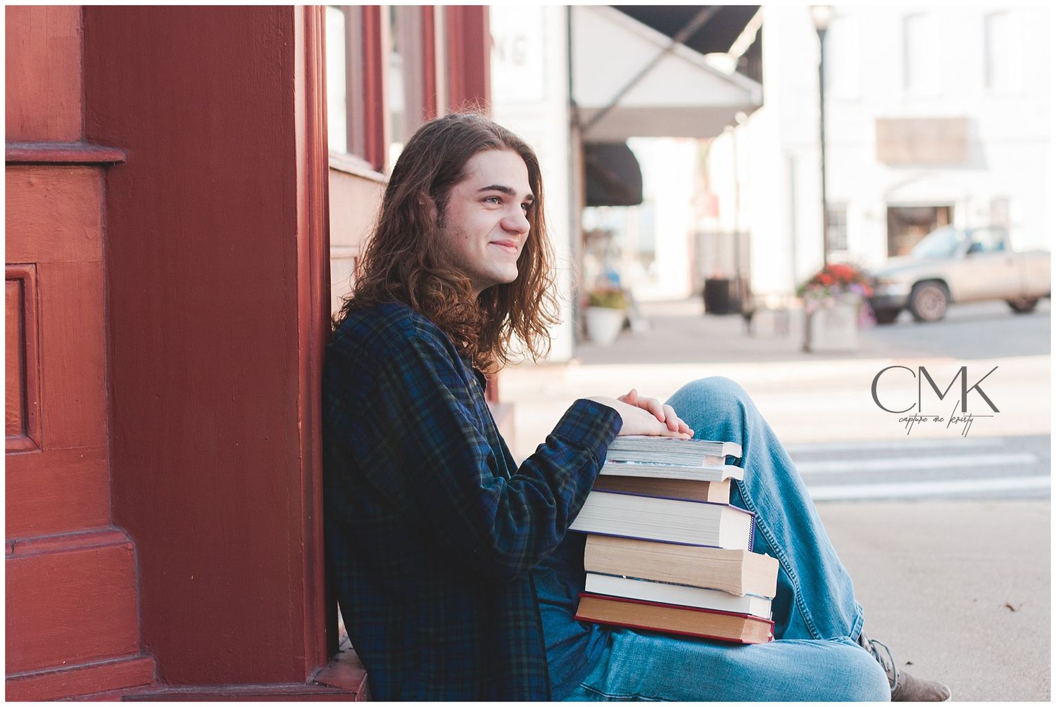 A person with long hair sits outdoors holding a stack of books against a red brick wall.