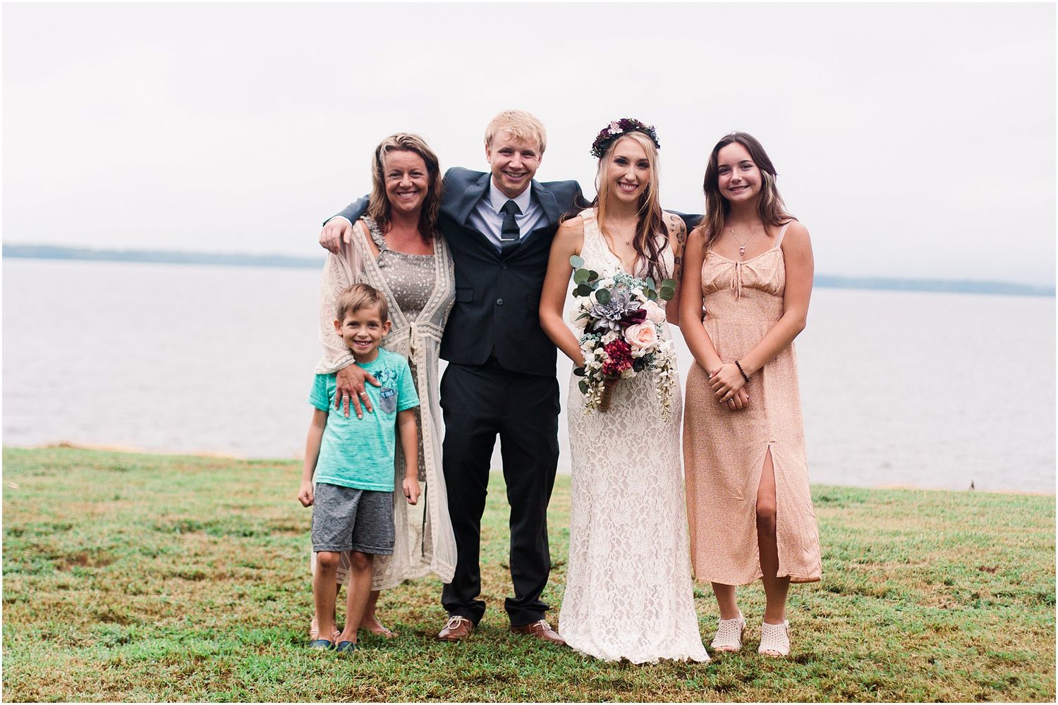 A wedding party poses together on grassy waterfront area in formal attire.