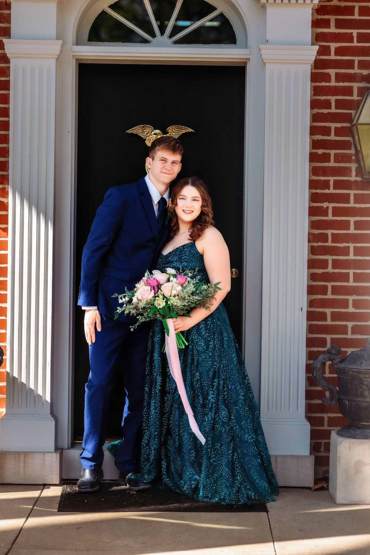 A couple in formal attire poses in front of a brick building with white doorway trim, holding a pink floral bouquet.