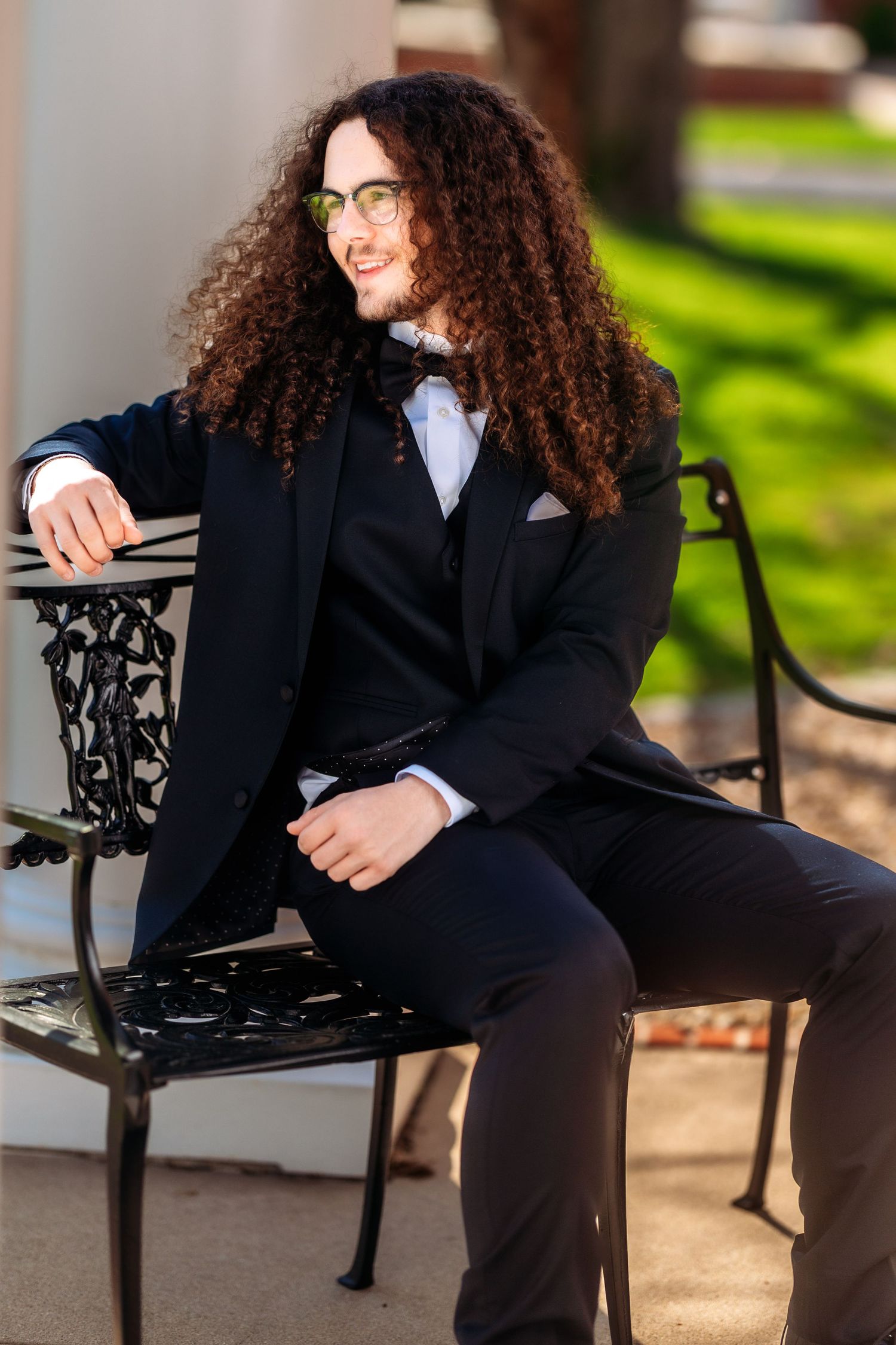 Elegant formal portrait of someone with long curly dark hair wearing a black suit sitting on an ornate bench outdoors.