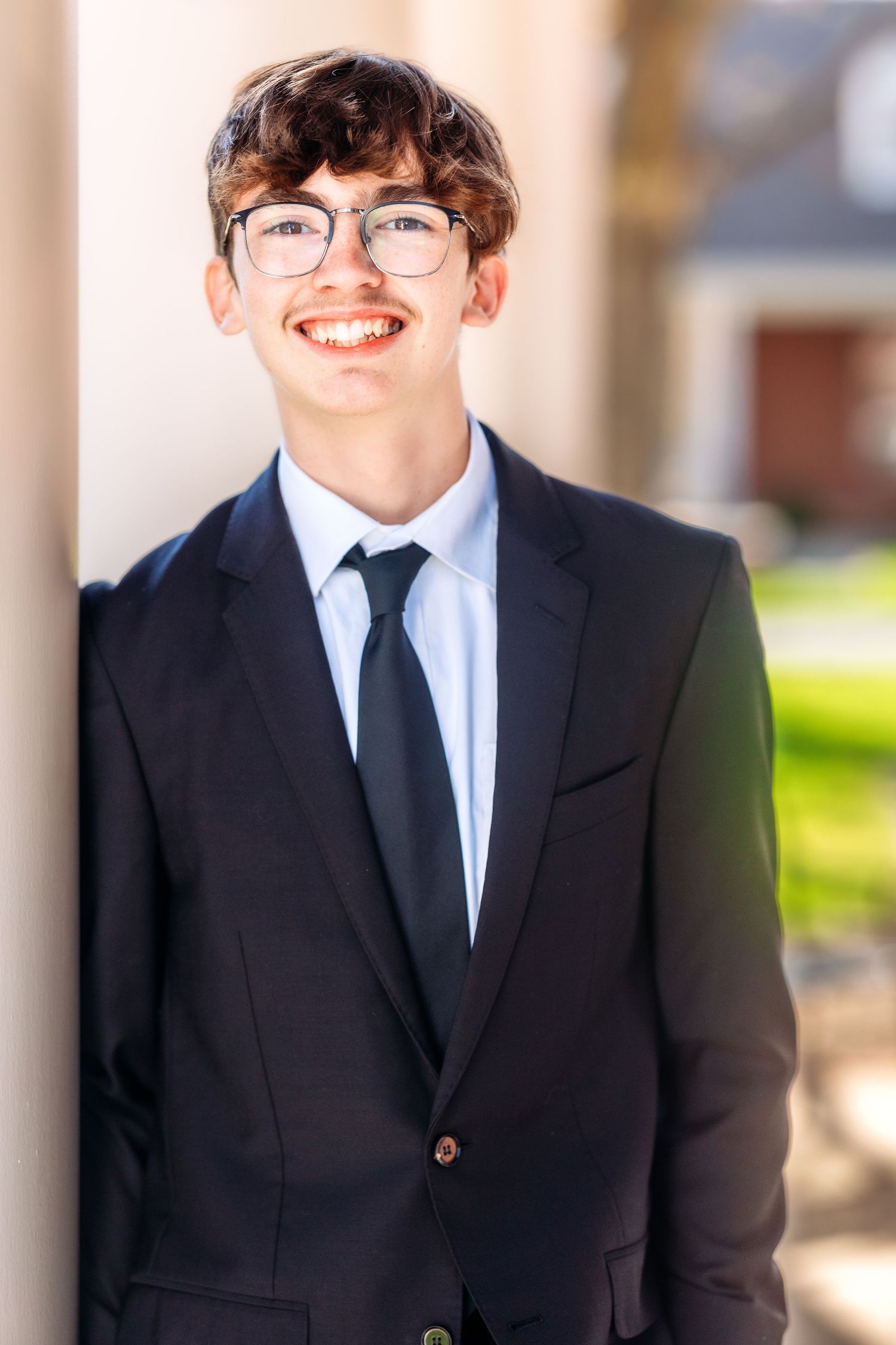 A person wearing a black suit and tie smiles warmly while standing outdoors in natural lighting.