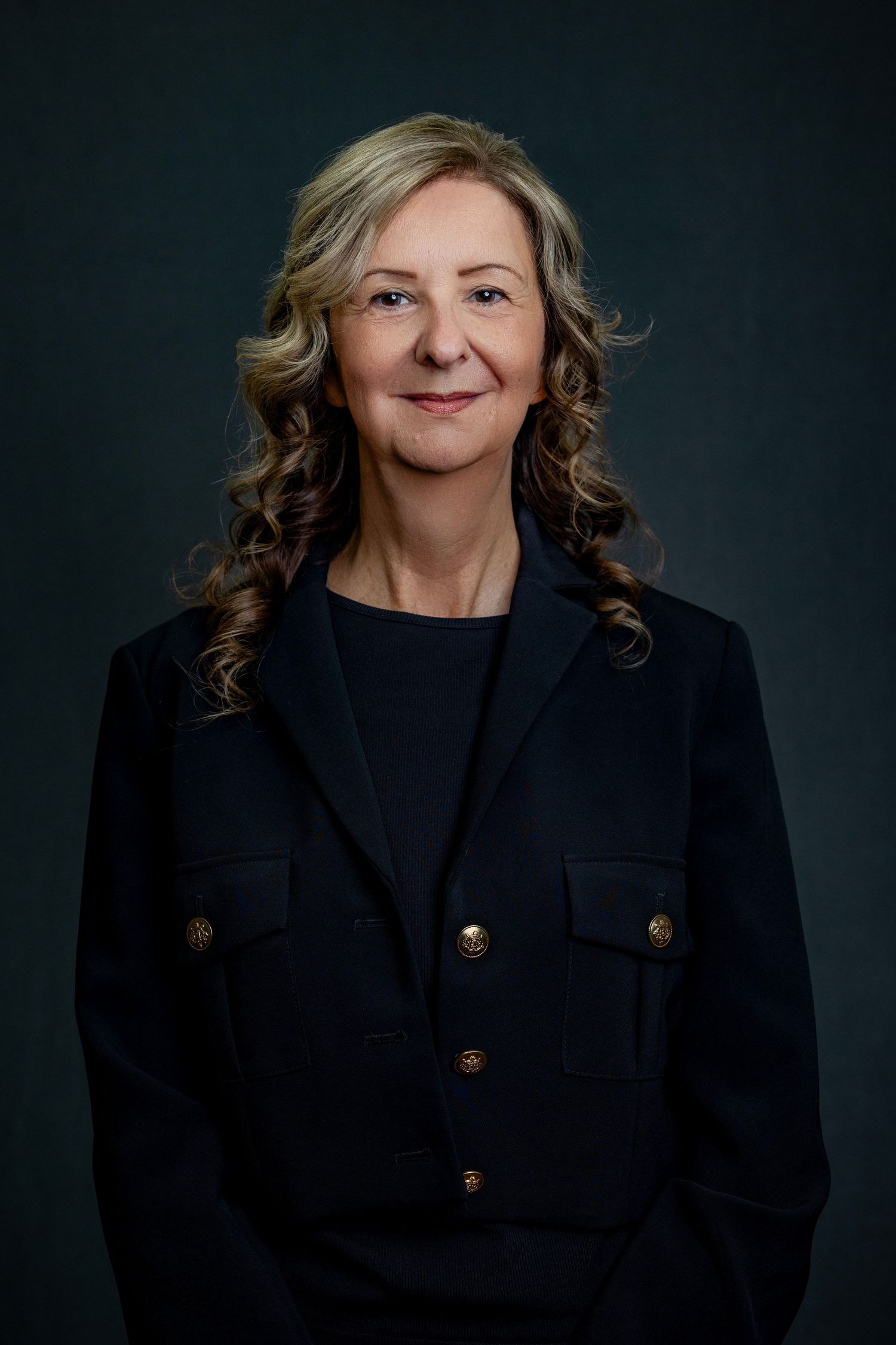 Professional headshot of woman wearing black blazer with gold buttons against dark background.