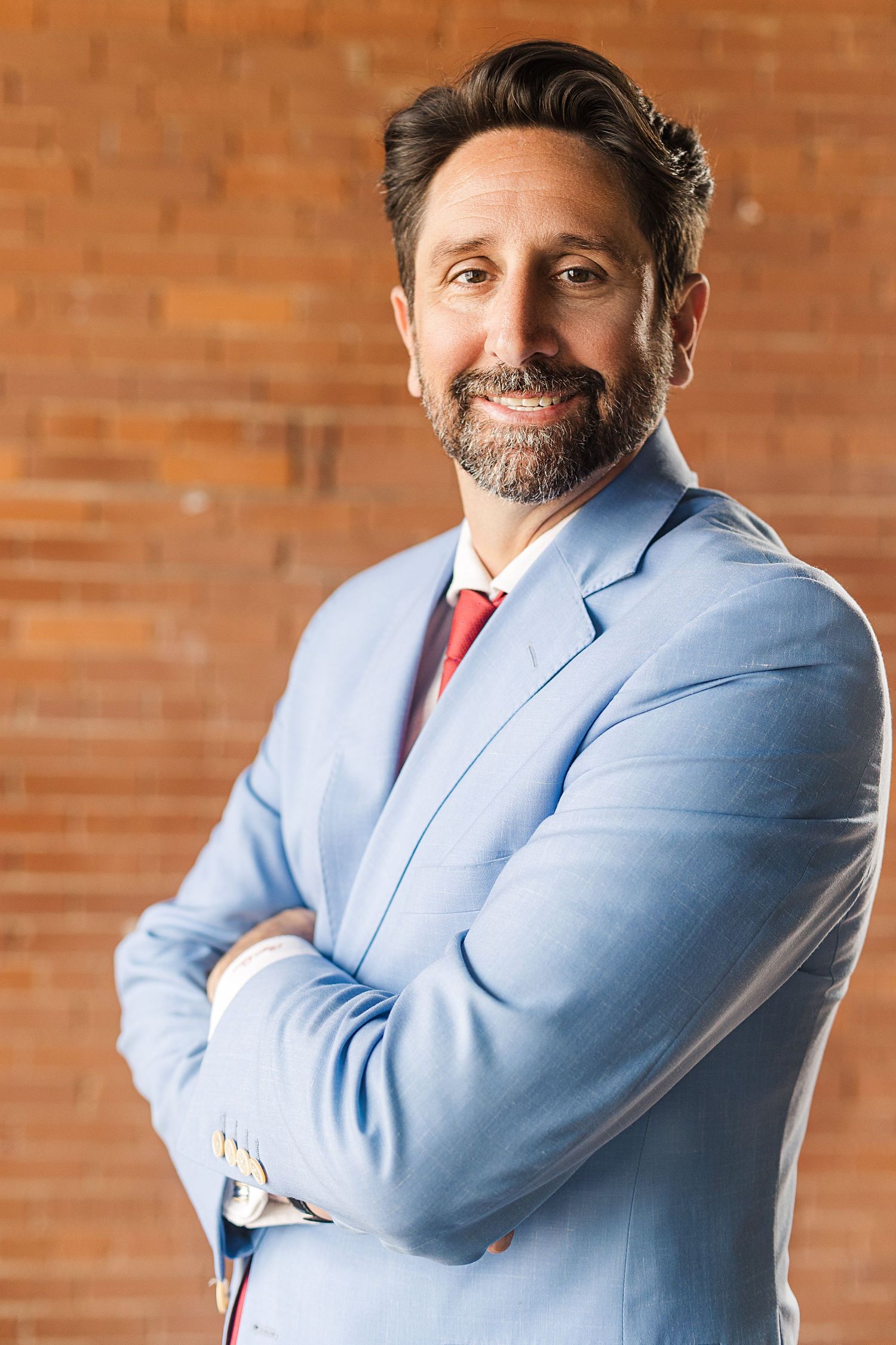 Professional portrait of a person in light blue suit with red tie against brick wall.