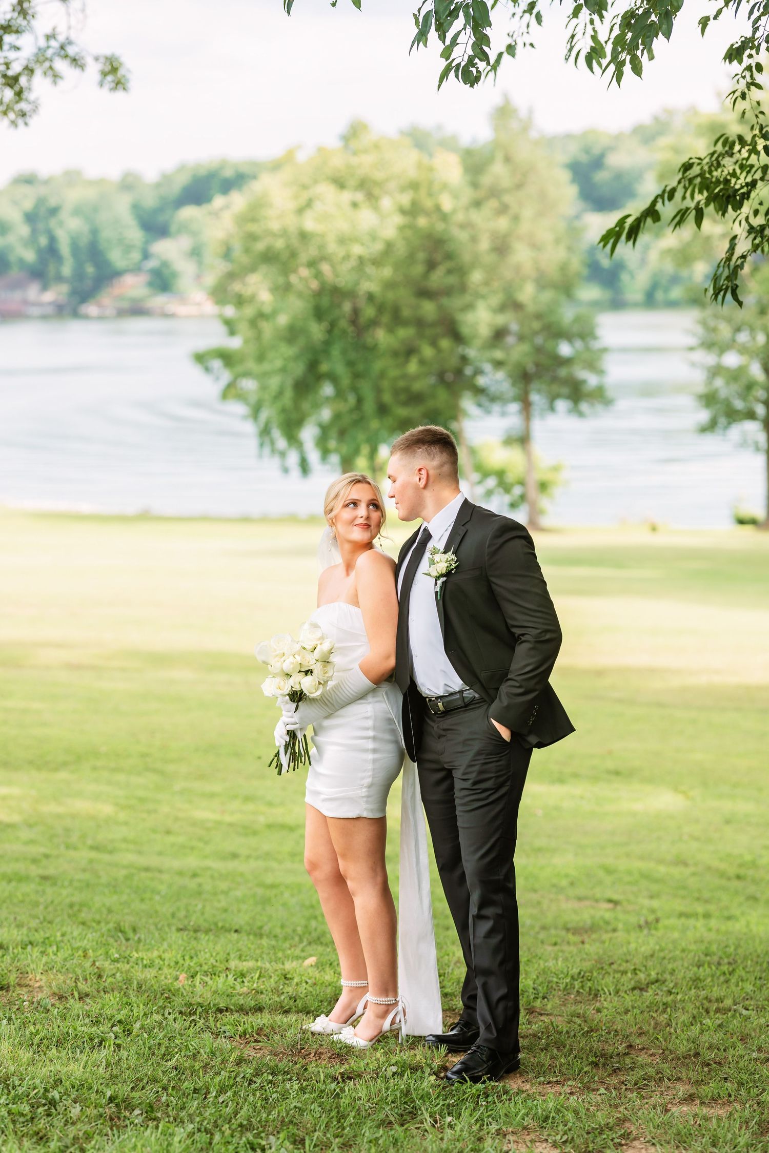 Wedding couple posing in formal attire on grassy lawn with lake view in background.